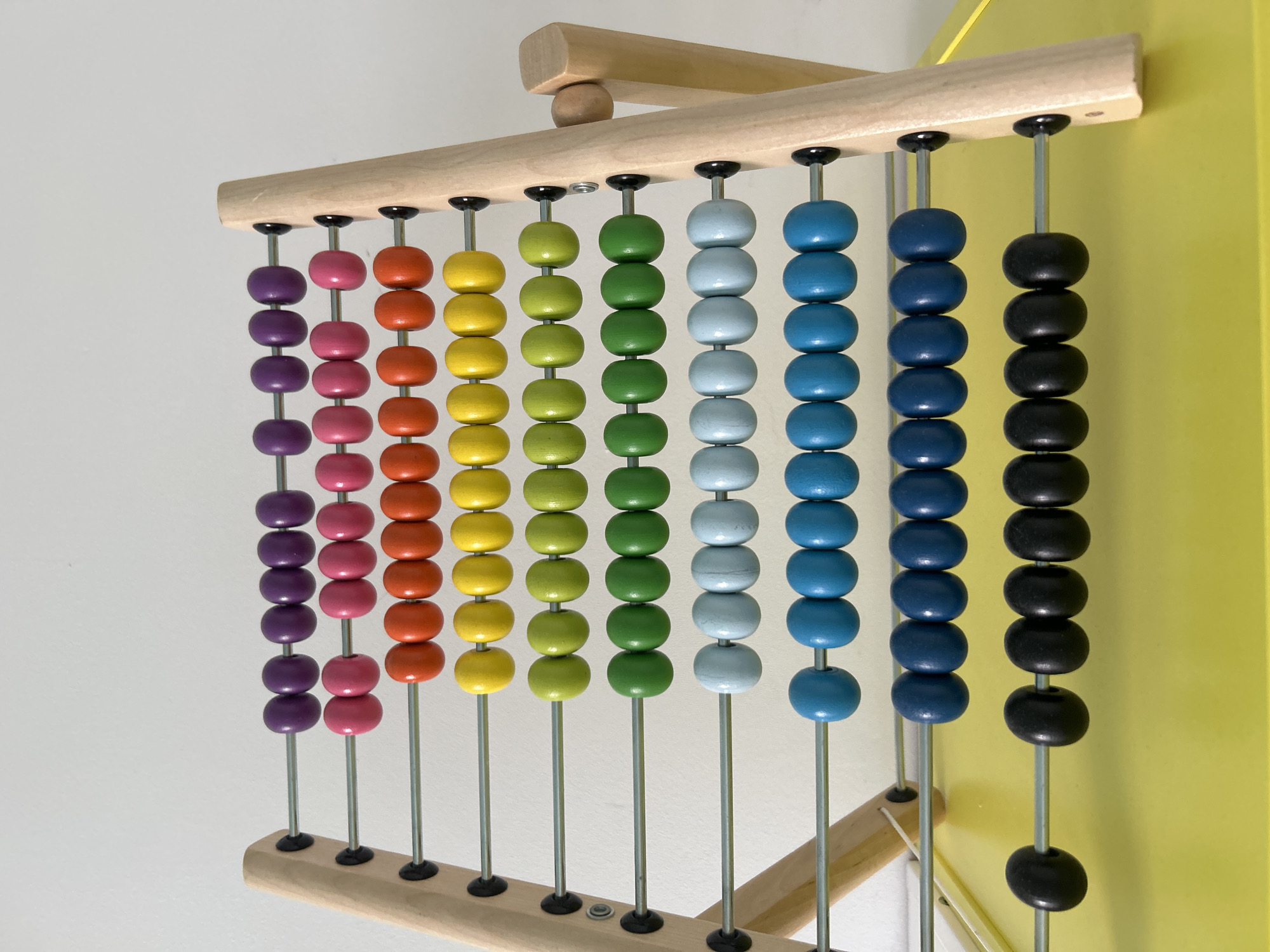 A close-up shot shows a wooden abacus with ten rows of ten beads each, arranged on a yellow surface against a plain white background. The beads are colored in sequence: purple, pink, orange, yellow, lime green, forest green, light blue, medium blue, dark blue, and black. Most of the beads are on the left side of the rods, with a few scattered across the different rows. The abacus is placed on a bright yellow surface, likely a table or tray. The lighting is bright and even, casting minimal shadows. There are no people or animals visible in the image, nor is there any indication of the city of Khimki, Russia.