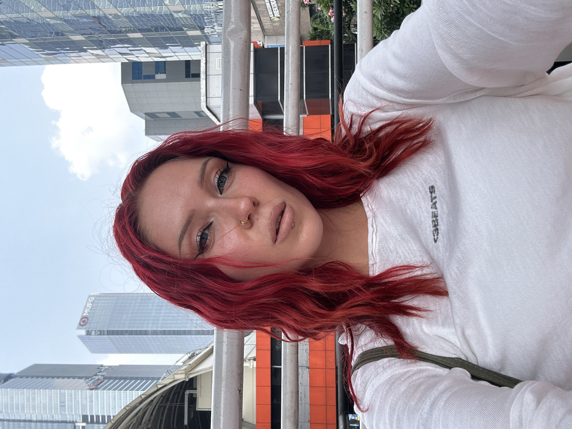 In a medium shot, a young woman with vibrant red hair and distinctive makeup, including dramatic eyeliner and lip piercings, looks directly at the camera. She is wearing a white long-sleeved shirt with a subtle logo. The background features modern urban architecture, including tall skyscrapers. One building in the distance displays the "MUFG" logo. This scene is situated in South Jakarta, Indonesia, specifically near the Directorate General of Immigration building, indicated by signage in the background. The overall environment appears to be an outdoor public space with a railing in the foreground.