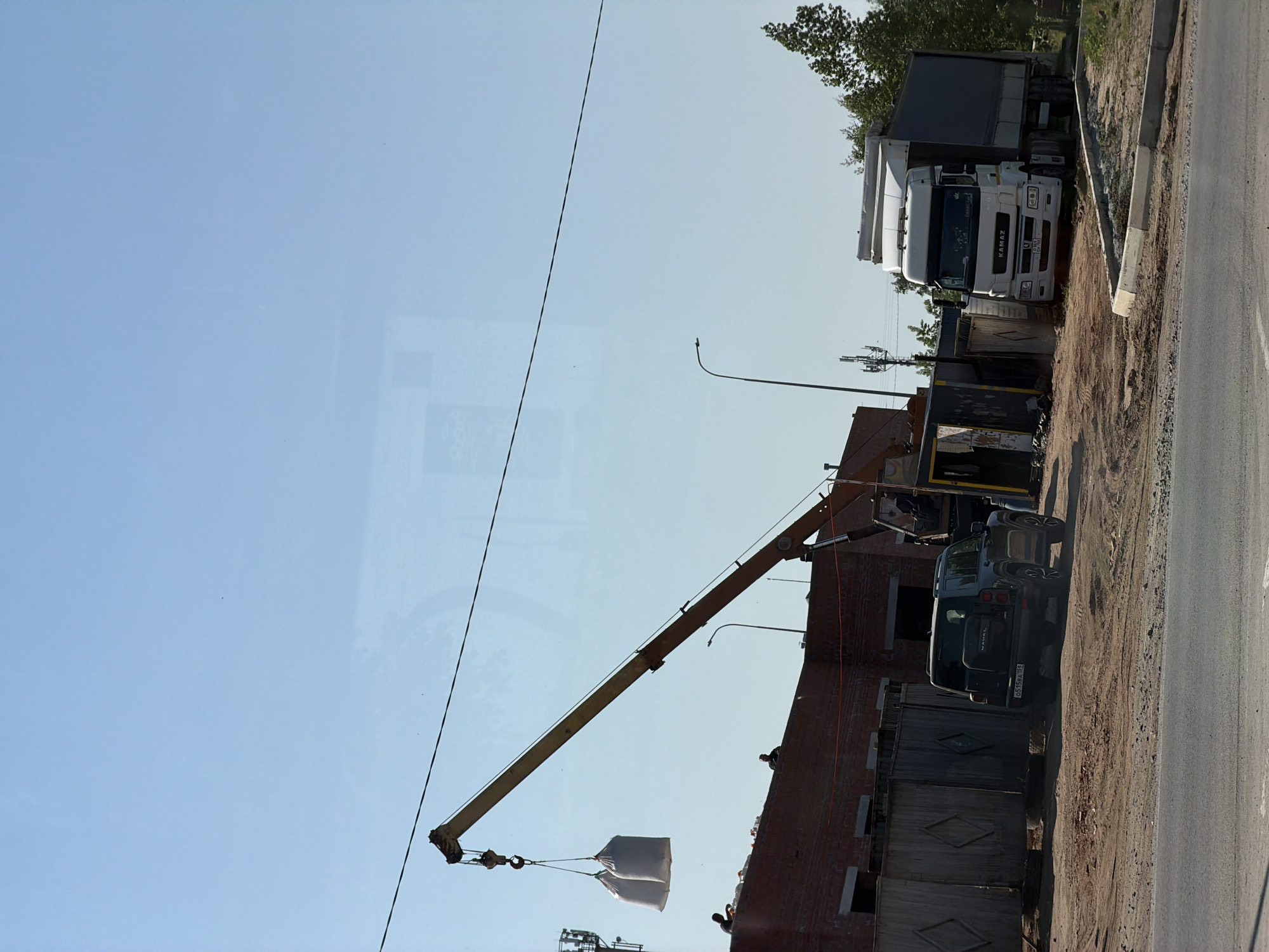 The image shows a construction site in Chaikovsky, Russia. A crane is lifting two large bags, possibly containing construction materials, onto the roof of a partially constructed brick building. A dark-colored HAVAL SUV is parked adjacent to the building. A KAMAZ truck is parked nearby, suggesting it might be involved in the construction project. The ground is dirt and gravel, and a paved road is adjacent to the construction site. Overhead wires and lampposts are visible. Several workers can be seen on the roof of the building. The sky is clear and blue.