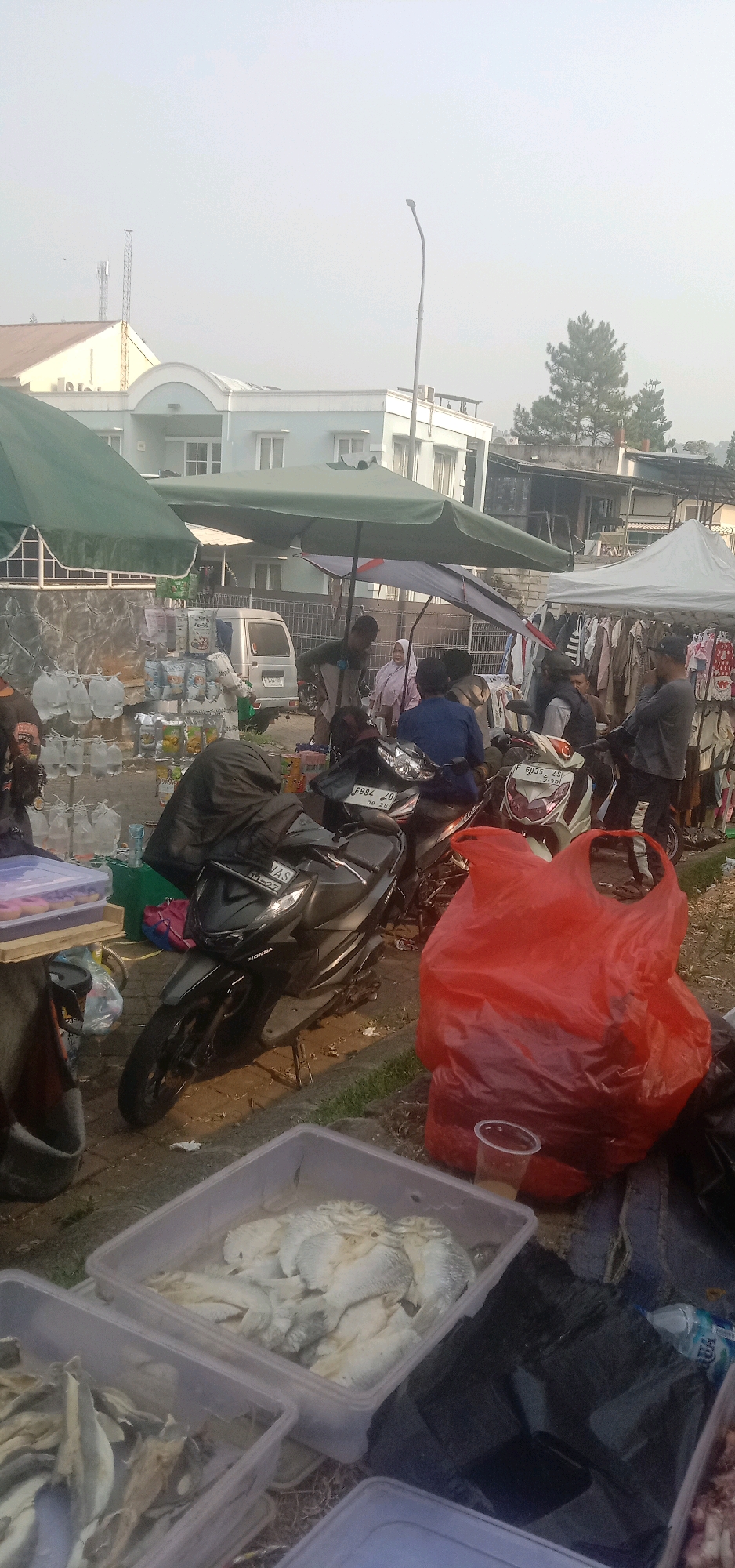 The image depicts an outdoor market scene in Sukanagalih, Indonesia. In the foreground, several translucent plastic containers hold what appears to be dried fish. To the right of these containers, a large, crinkled red plastic bag is prominent, alongside a clear plastic cup. Behind these items, motorcycles are parked, with one black scooter in the mid-foreground displaying the license plate "F 6884 ZD". Several people are visible in the background, some standing and some seated on the motorcycles. They appear to be interacting within the market environment.
The market is set up with various stalls covered by umbrellas and canopies. One stall features racks displaying clothing for sale. Another stall is laden with packaged goods, some of which are stacked high. Several rows of clear plastic bags filled with water are also visible, likely for sale. A silver van is parked behind one of the stalls. In the upper portion of the image, buildings are visible, including what appears to be a multi-story structure with windows and a light blue facade, as well as a smaller building with a brown roof. Trees and a communication tower are also present in the background, suggesting a semi-urban or suburban setting. The sky is overcast and hazy, indicating subdued lighting conditions. The ground surface appears to be paved with interlocking blocks in some areas, transitioning to a dirt or gravel path.