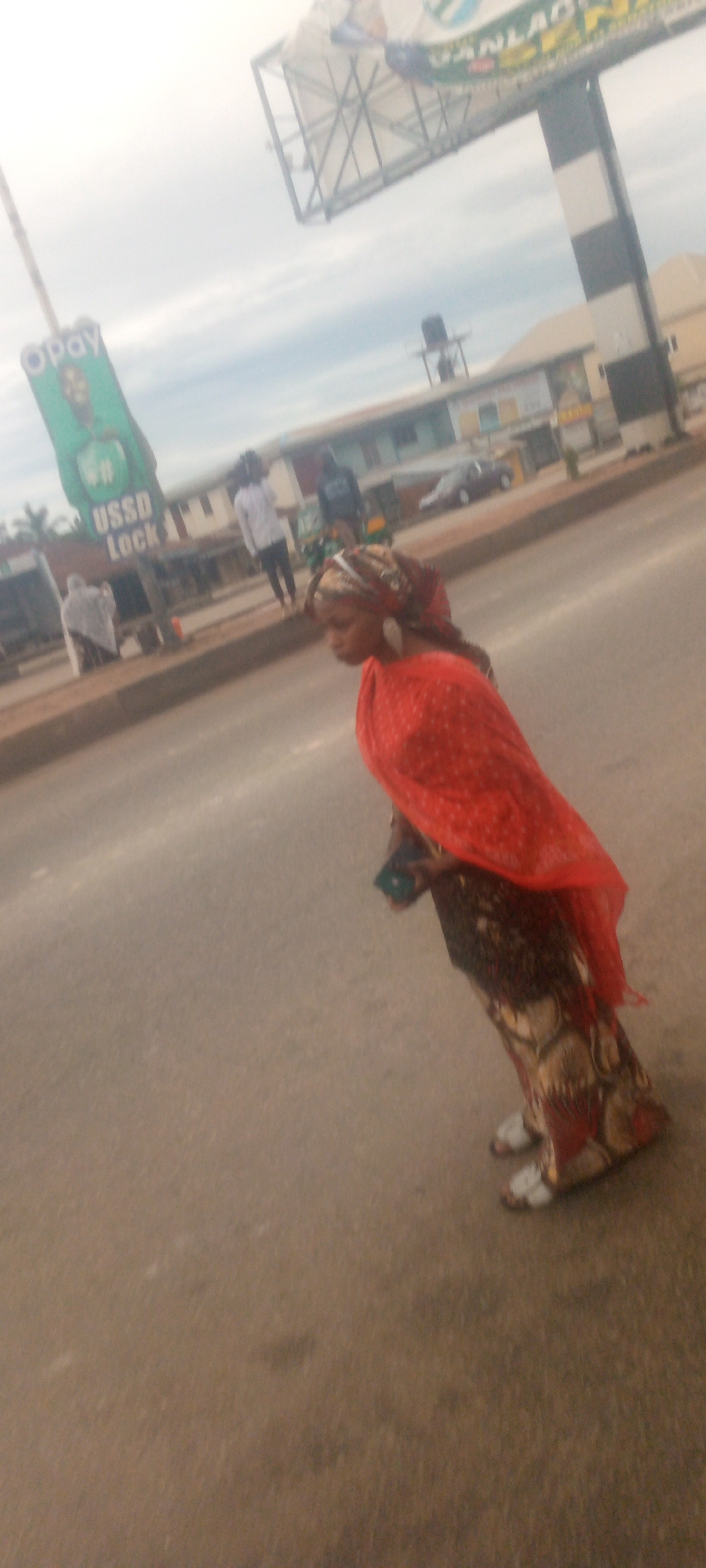 A low-angle, slightly blurry, medium shot captures a woman walking on a paved road in an urban environment. She is wearing a vibrant red headscarf and a patterned dress, with white sandals on her feet. She is looking down and to her right, and holding a dark-colored object, possibly a phone, in her left hand. In the background, a billboard advertising "OPAY USSD Lock" is visible on the left, with the "OPAY" logo prominently displayed. Further in the background, buildings line the street, and other pedestrians and a vehicle are present. The sky is overcast, suggesting a cloudy day. The overall scene depicts a moment of everyday life on a street in an unnamed city.