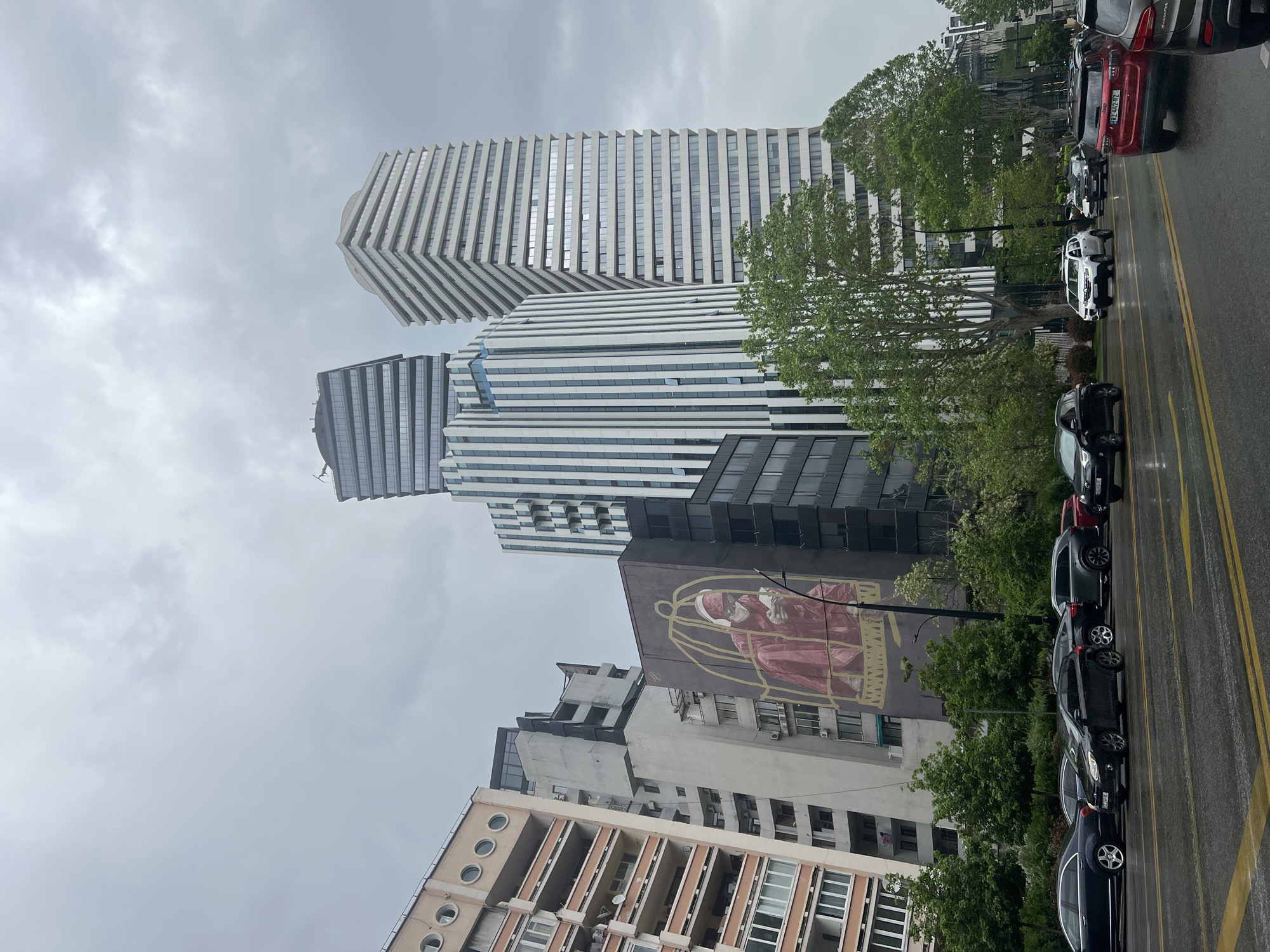 The image shows a street scene in Tbilisi, Georgia, featuring a row of parked cars alongside the road with yellow lane markings. The sky is overcast. Buildings of varied architectural styles rise in the background. Two contemporary high-rise buildings with horizontally striped façades are prominent. A mural depicting a figure inside a cage adorns the side of another building.