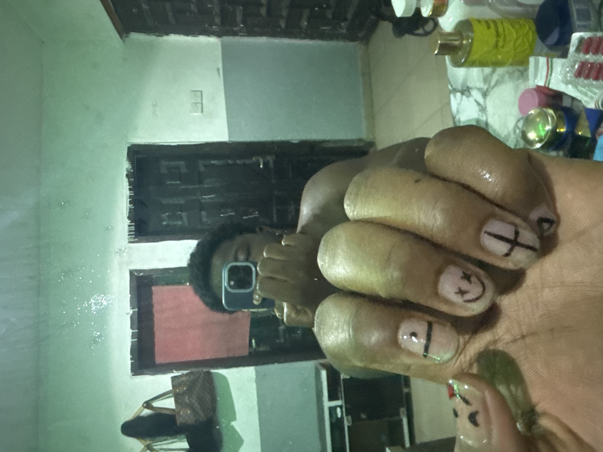 A person is taking a selfie in a mirror. The person has a dark complexion and short, dark hair. The phone being used has a light blue or gray case. The person's hand, with painted fingernails, is prominent in the foreground. The fingernails are a pale color with black designs. The background reveals a room with light green walls and a dark wooden door. Various items, including bottles and containers, are on a surface to the right. A purse or bag is visible in the lower left corner. The reflection is likely inside a domestic residence. There are no clear landmarks to establish the location as Gbongan, Nigeria.