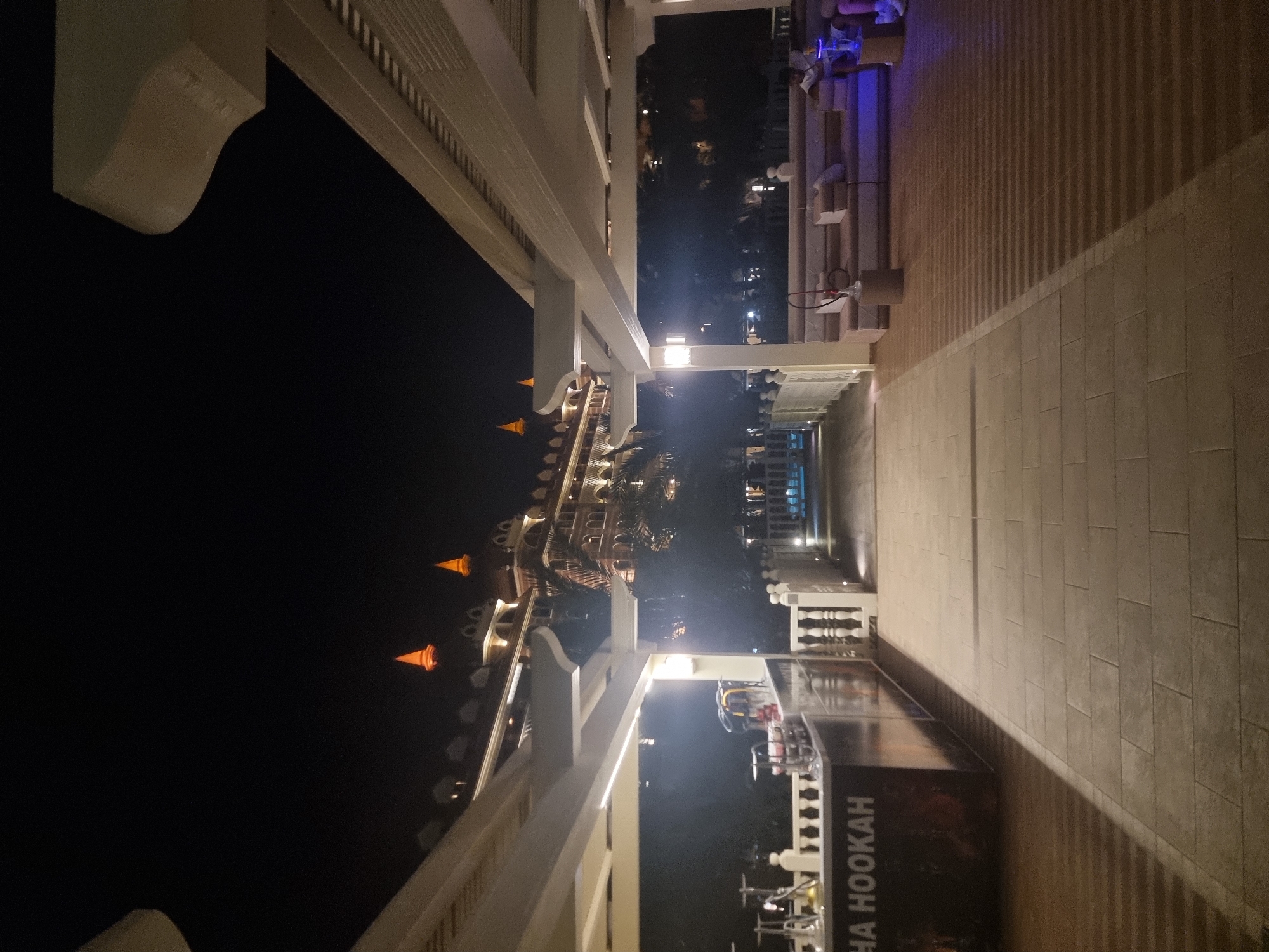 The image depicts a nighttime scene at a resort, likely in Manavgat, Turkey. In the foreground, a walkway paved with large rectangular tiles extends into the distance. To the left, a bar area is visible, with the text "HA HOOKAH" emblazoned on it. Several shisha pipes are displayed on the counter. Beyond the bar, a white railing lines a walkway.
In the mid-ground, a pathway leads towards a building. The building itself is illuminated, with prominent pointed roof structures that glow orange. It appears to be a large resort or hotel with multiple stories and ornate architecture. Palm trees are visible in front of the building.
To the right of the walkway, a covered seating area with low sofas and cushions is set up. Several shisha pipes are placed on low tables in front of the sofas. Two individuals are seated on the sofas, one appearing to be a young person. The seating area is illuminated with blue ambient lighting. The overall environment suggests a recreational area at a hotel or resort, likely offering hookah services. The lighting creates a warm and inviting atmosphere despite the darkness.