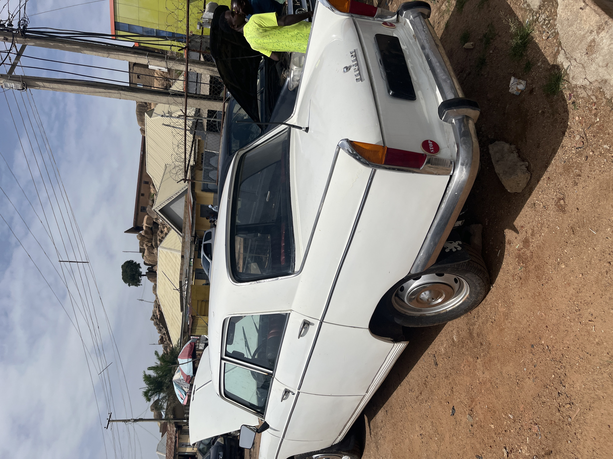 A white Peugeot sedan with its hood open is parked on a dirt lot adjacent to a street in Jos, Nigeria. The sedan is of an older model, evident by its boxy frame and chrome bumpers. A man wearing a bright green shirt stands near the front of the vehicle with the hood raised, apparently examining the engine. The surroundings include utility poles, barbed wire, and buildings of varying heights. The ground is predominantly brown dirt, typical of the local soil.