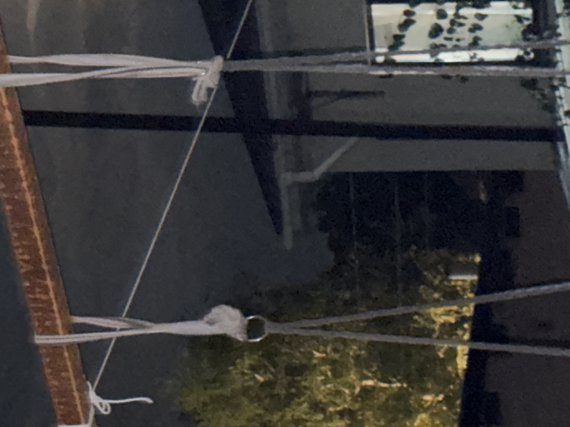 The image depicts a close-up view of ropes suspended from a rusty metal beam. The ropes are white and grey and appear to be made of fabric. One rope is tied to the beam with a knot, and a metal ring is attached to another rope. In the background, a tree with green foliage is visible, along with a dark, out-of-focus building. The lighting suggests it is either dusk or nighttime. The scene is indoors or under a covered structure, possibly a porch or carport, given the presence of the beam and the hint of a building's roofline. There are no people or animals visible in the image. The location in Vostok, Kyrgyzstan is not discernable from the visual information provided.