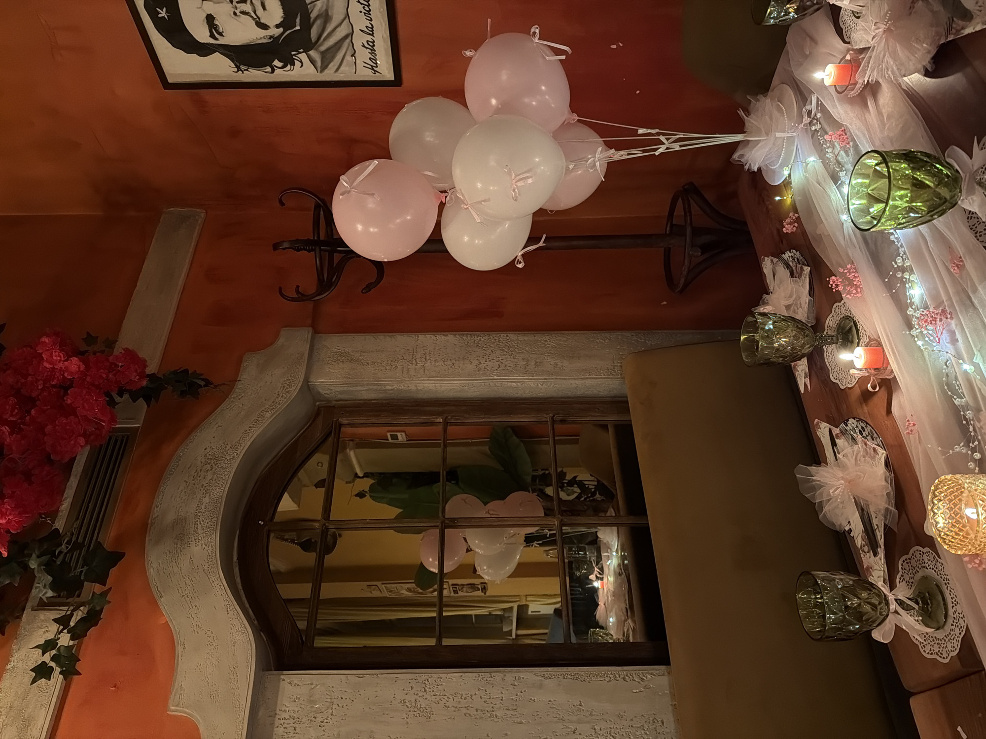 The image depicts an interior space, possibly a restaurant or event venue, with a focus on decorations. A cluster of pink and white balloons is tied to a wrought iron stand. Reflected in a nearby window are the balloons and the decor. A table is set with ornate glassware, candles, and sheer white fabric, suggesting a celebratory setup. The wall is painted in a terracotta tone, with an architectural detail featuring a decorative arch. A framed poster of Che Guevara is visible on the upper portion of the wall. Based on the decor style, it could be a themed restaurant and there is no information to support or deny it is from Sofia, Bulgaria.