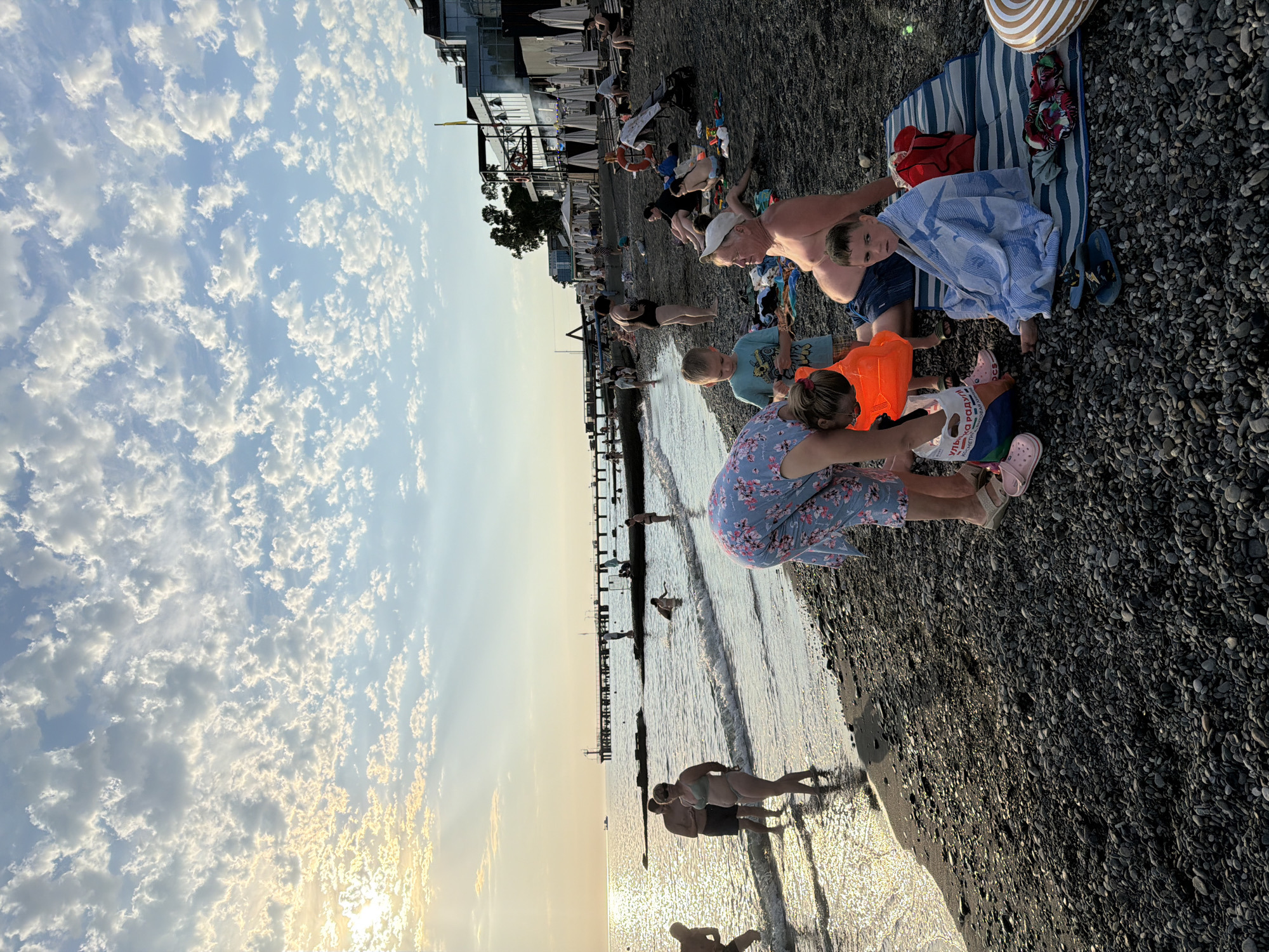 The image depicts a crowded beach scene in Sochi, Russia, during what appears to be the late afternoon or early evening. The sky is filled with scattered, puffy white clouds against a pale blue backdrop, with the sun setting on the left side of the frame, casting a warm glow over the water and creating shimmering reflections. The beach itself is composed of dark gray pebbles.
In the foreground, several people are engaged in various activities. A woman in a floral dress is bending down, tending to a young child wearing an orange life vest. Beside them, a young boy in a blue t-shirt is looking down at something in his hands. An older man with a white cap is sitting on a striped blanket, partially covered by a blue and white striped towel. Another young boy is sitting on the blanket, also wrapped in a towel. Various beach items, including bags, towels, and flip-flops, are scattered around the people.
Further back, a group of people are standing in the shallow water, with waves gently lapping at the shore. Some are wading, while others appear to be interacting. In the middle ground, a pier extends into the sea, with a few figures visible on it. In the background, a line of buildings, likely hotels or restaurants, can be seen along the coastline, some with large windows and balconies. Umbrellas are set up in front of some of the establishments. A small boat is visible on the horizon. The overall atmosphere is one of a busy public beach during a pleasant weather day.