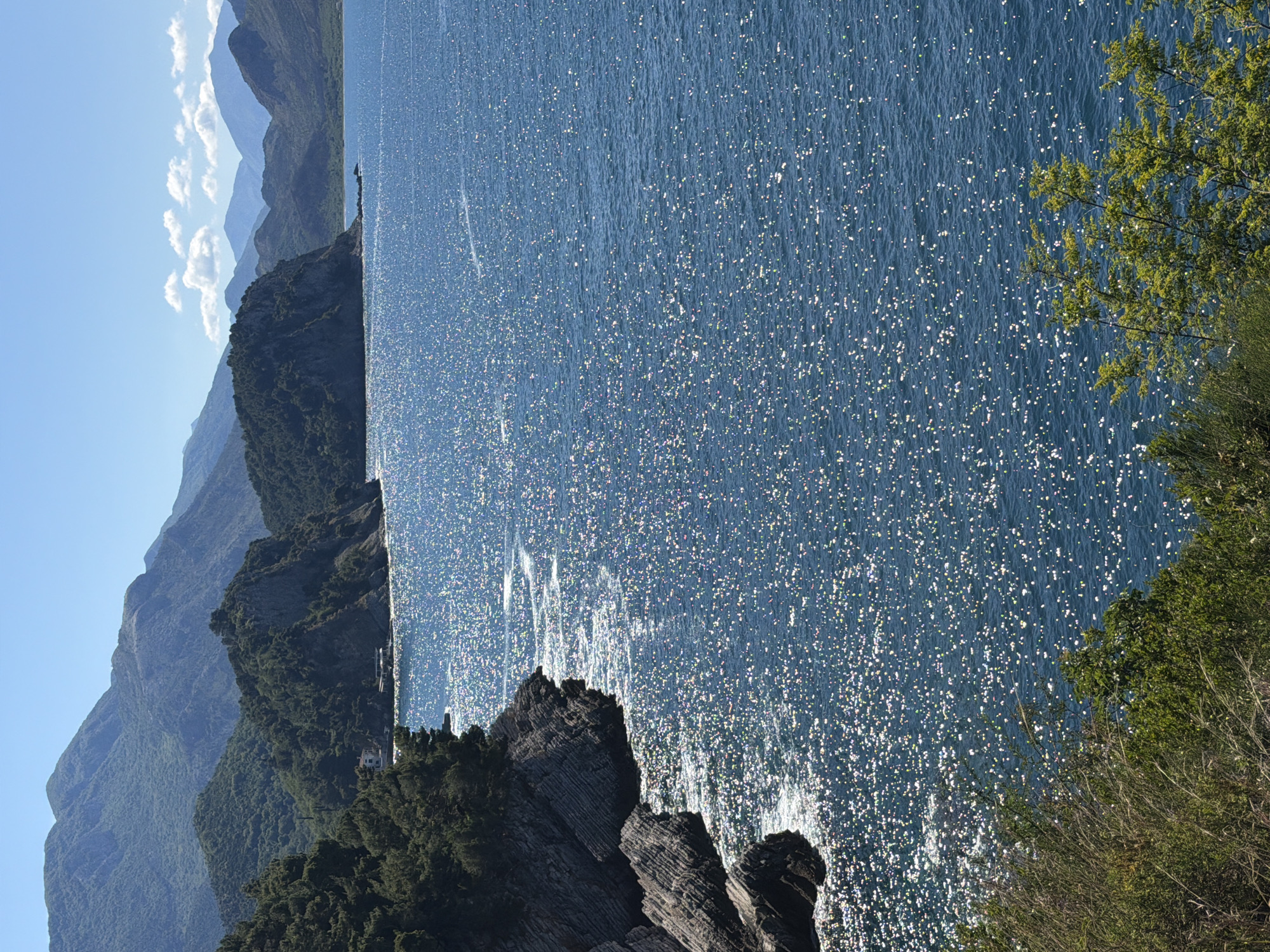 The image shows a body of water, possibly the Adriatic Sea, glistening with sunlight. In the background, layers of mountains recede into the distance, their peaks obscured by haze and clouds. In the lower left, a rocky coastline is visible, with sparse vegetation clinging to the rocks. Green foliage frames the right side of the image. Given the prompt, the location is likely Petrovac na Moru, Montenegro. There are no people visible in the image.