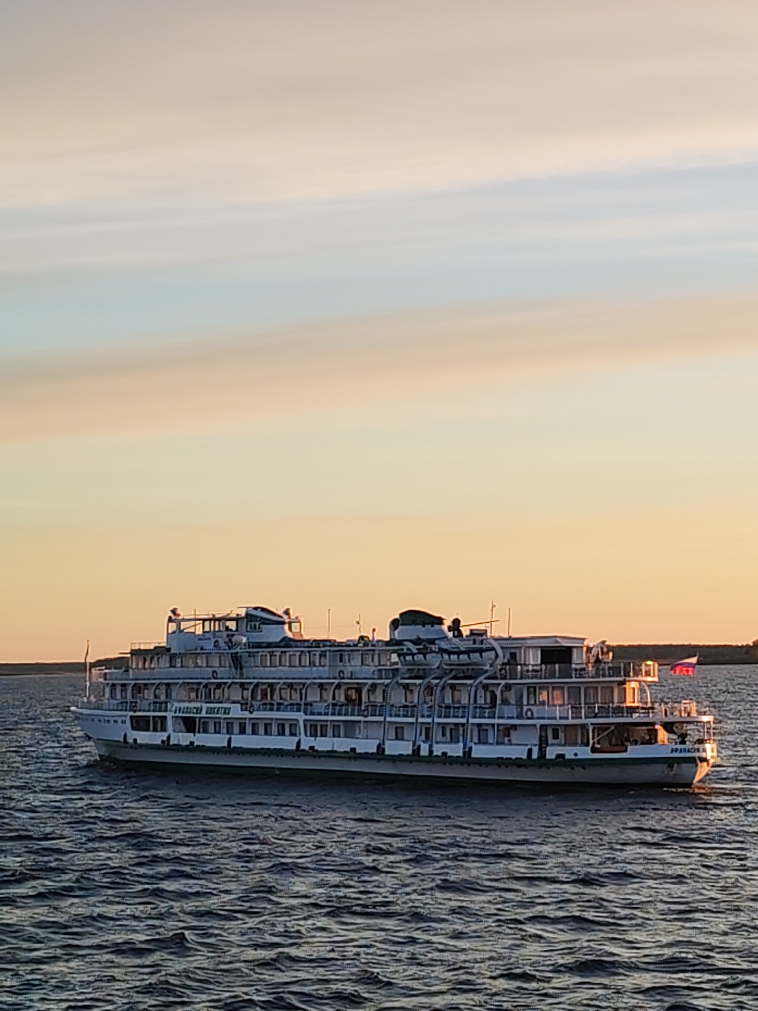 A large, white, multi-level cruise ship, the "Anatoly Lyapin," sails on a body of water, likely a river or lake, near Mikhalevo, Russia. The ship features multiple decks with windows and railings and a Russian flag is on the stern. The sky shows shades of light blue, yellow, and orange, suggesting it is either sunrise or sunset. The water is dark with visible ripples. The shoreline is faintly visible in the background.