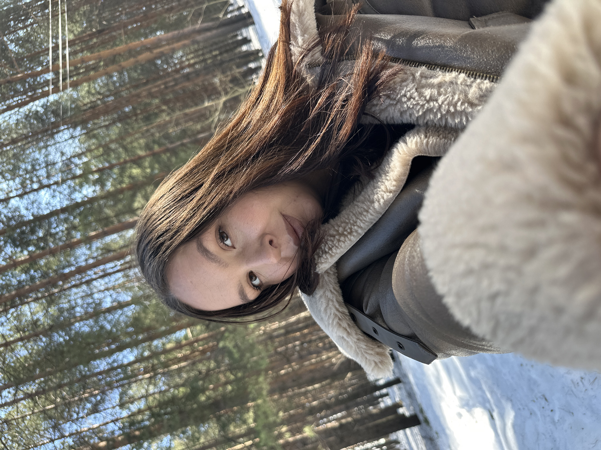 The image depicts an adult female taking a selfie in a snowy pine forest, located in Chaikovsky, Russia. The subject, positioned in the foreground and facing the viewer, has long dark brown hair, light brown eyes, and fair skin, with a neutral facial expression. She is wearing a brown aviator-style jacket featuring a prominent cream-colored shearling collar and cuff. Her right arm, covered by the jacket, is extended towards the camera, suggesting the selfie action. The environment consists of a dense arrangement of tall, slender pine trees with reddish-brown trunks. The ground is uniformly covered with white snow, exhibiting some disturbed areas or tracks. The scene is brightly illuminated by direct sunlight, creating distinct shadows. Two thin, dark, horizontal lines, consistent with utility wires, are visible in the upper right background above the tree line.
