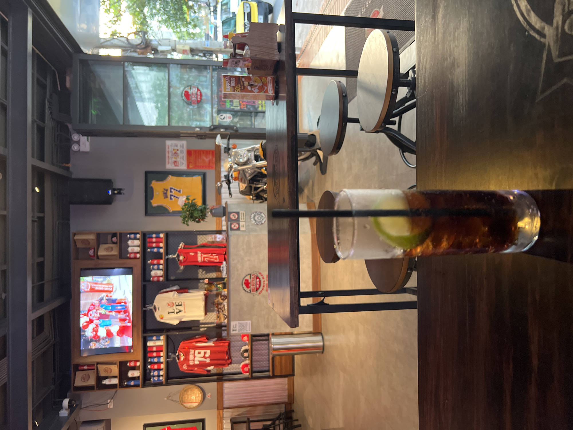 Here is a detailed description of the image:
**Setting:** The photo appears to have been taken inside a bar or restaurant, likely in Bắc Nha Trang, Vietnam, given the decor that includes sports memorabilia and the presence of the "Red Buffalo Hub" logo, a sports bar chain with locations in Vietnam.
**Objects and Environment:**
* **Interior:** The interior is decorated with sports jerseys and memorabilia displayed on shelves along a wall. A television screen is playing what appears to be a sports game.
* **Furniture:** The image focuses on a high table with stools. The table has a metallic frame with a wooden top.
* **Drink:** A glass of what seems to be a mixed drink with a lime wedge and a straw is on the table.
**People and Interactions:**
* There are no people visible in the image, the scene captures an empty bar interior.
**Subject:**
* The main subject is the drink placed on the counter of the bar.