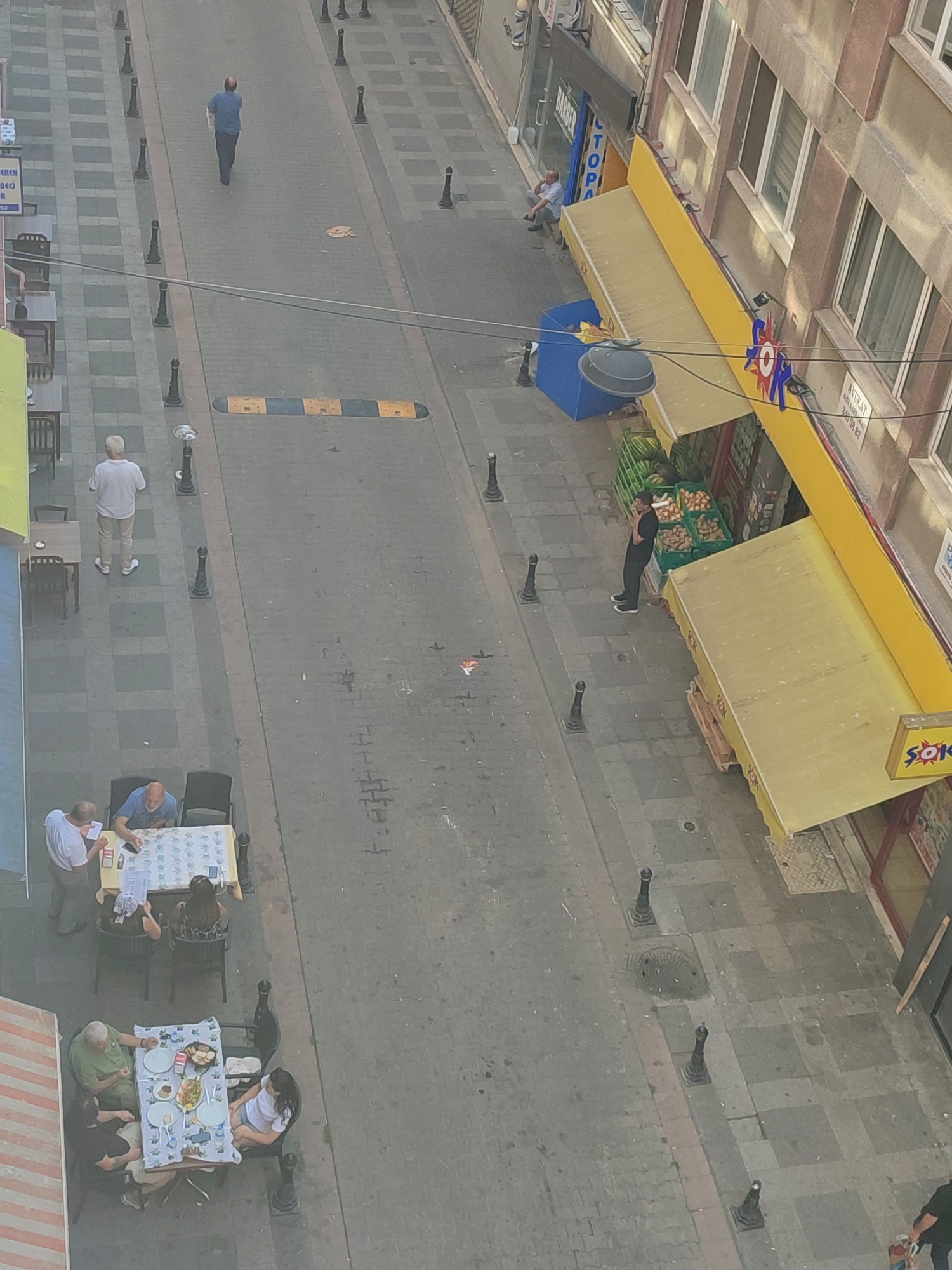 This aerial, oblique photograph depicts a street scene in Kadıköy, Turkey. The perspective is high, looking down at a pedestrianized street paved with grey interlocking stones, interspersed with checkered grey and tan square patterns. The street is lined with businesses on the right side, featuring yellow awnings over storefronts. One storefront displays a red and blue starburst logo and text in Turkish, suggesting a market or store. Another awning has a yellow sign with the text "SOK" in red, indicating a likely supermarket.
Several individuals are visible in the image. On the left side of the street, two tables with chairs are set up outside a building. At one table, three people are seated, and another person is standing and appearing to interact with them. At a second table further down, four people are seated and engaged in what appears to be a meal. On the right side of the street, a man in dark clothing leans against the wall of a building near a display of produce, including watermelons and other fruits. Further up the street, a man in a blue shirt walks away from the camera carrying a white object. Near the storefronts, another man is seated on the curb.
Various street furniture and objects are present, including black bollards lining the street, a blue dumpster, and speed bumps across the roadway. The overall impression is of a bustling urban environment, likely a commercial area with outdoor dining and retail activity. The lighting suggests it is daytime.