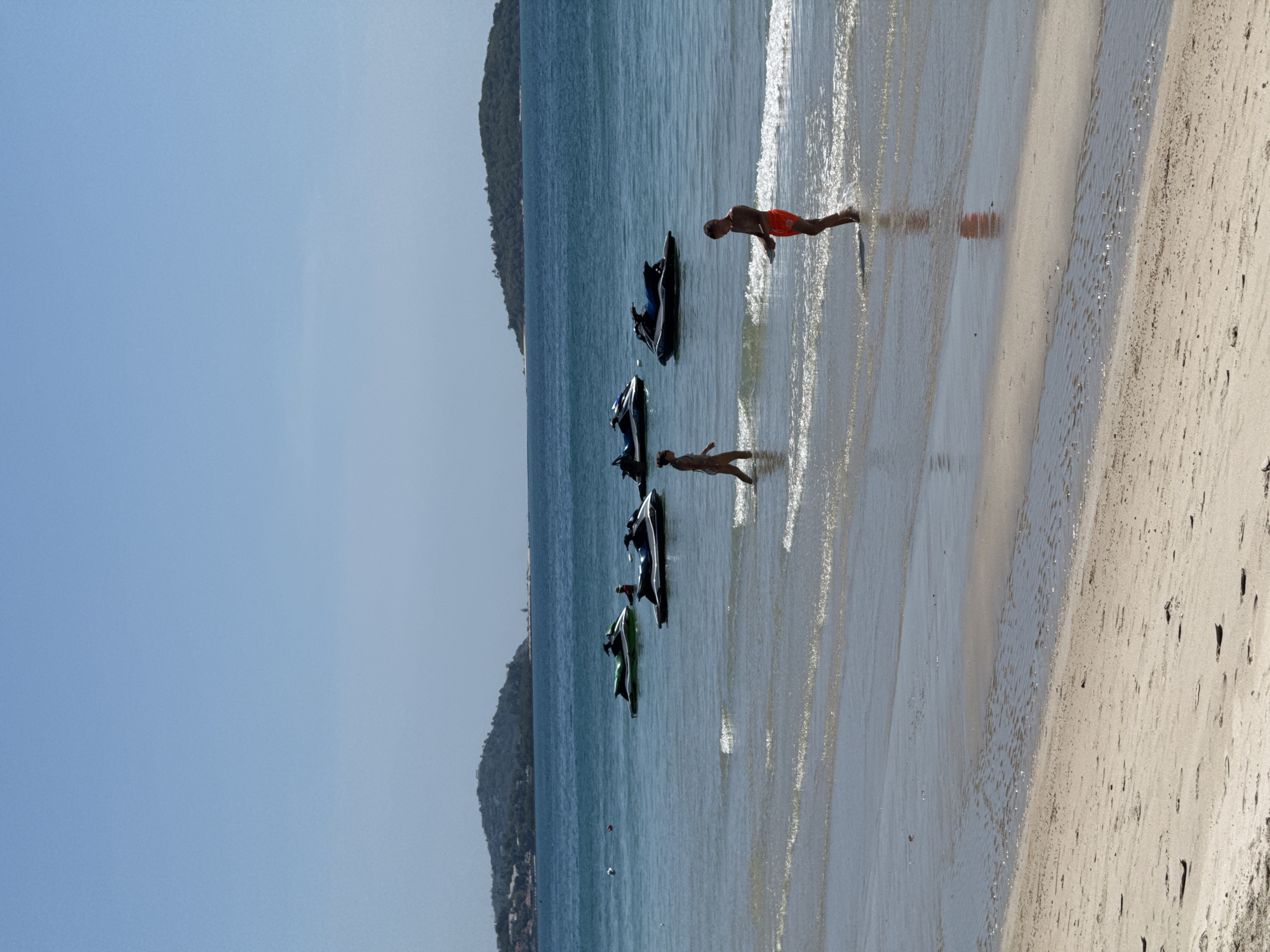 In the city of Ko Samui District, Thailand, a bright, sunny day unfolds with a sandy beach leading to the turquoise waters of the Gulf of Thailand. Several jet skis are scattered across the calm sea, some with people on them, and others anchored. In the foreground, two individuals are wading in the shallow water. A person in a bikini is walking towards the left, while another person in red shorts is standing with their back to the camera, looking at the water. In the background, lush green islands rise from the horizon, with a few buildings visible on the slopes of the island to the left. The sky is a clear, pale blue.