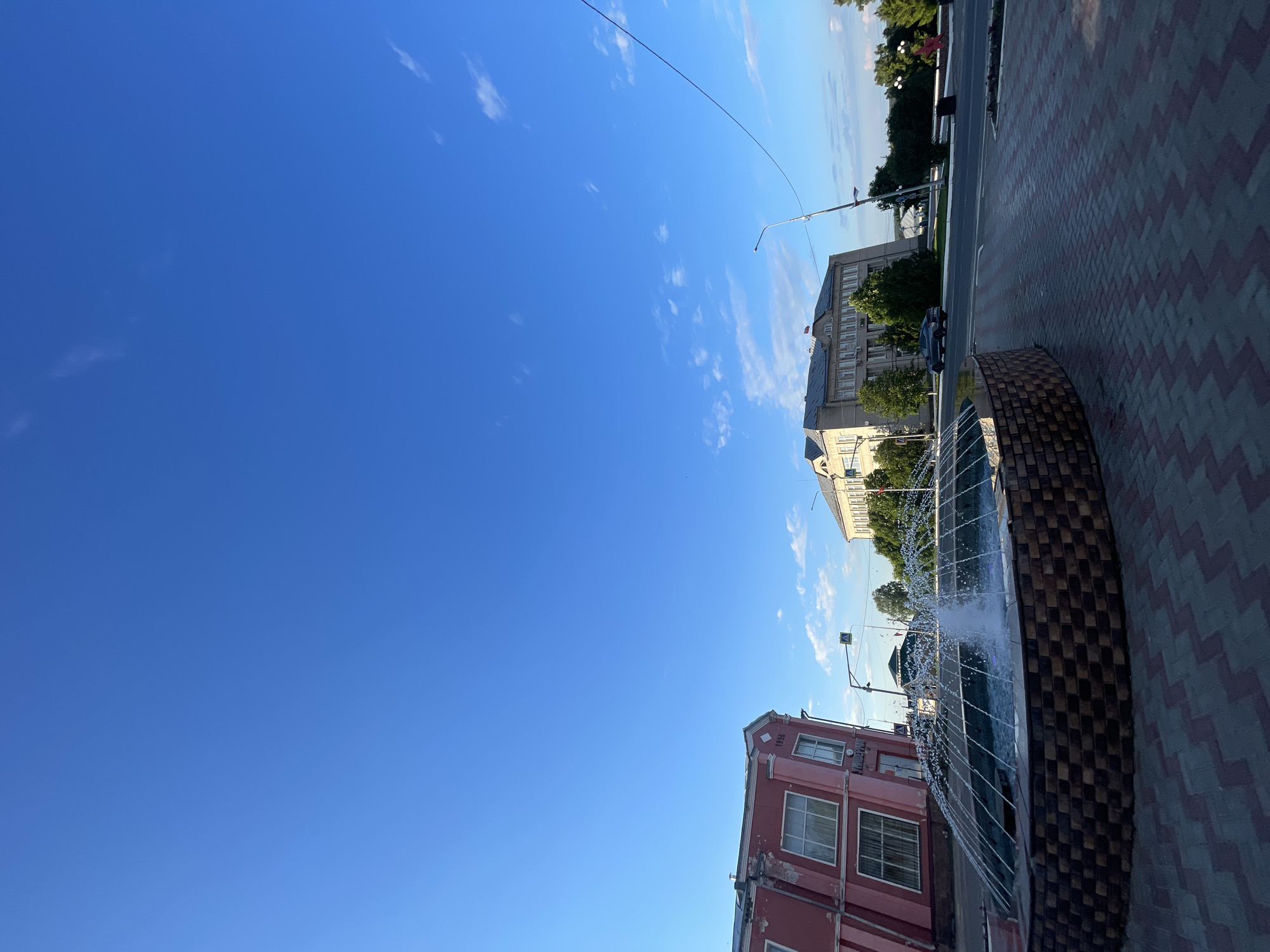 The image depicts a street scene in the city of Krasnye Baki, Russia, on a clear day with a bright blue sky and scattered white clouds. In the foreground, a circular fountain with a brick-patterned base is actively spraying water, creating a misty arc. The ground around the fountain is paved with interlocking brick pavers in a zig-zag pattern of red and grey.
To the left of the fountain is a two-story building, painted a faded pink, with white trim around its windows. The building appears to be older, with some visible wear and tear on its facade. Above one of the windows, the year "1956" is inscribed.
Across the street from the pink building is a larger, lighter-colored building with a more modern architectural style. It has multiple windows and a pitched roof. In front of this building, there are several mature green trees.
A paved road runs in front of both buildings and the fountain. A dark-colored car is visible driving on the road. Traffic lights and street lamps are present along the street. The overall environment suggests a public space within the city, likely a town square or a central street intersection. The presence of the fountain and well-maintained paving indicates an area designed for public use and possibly recreational value.