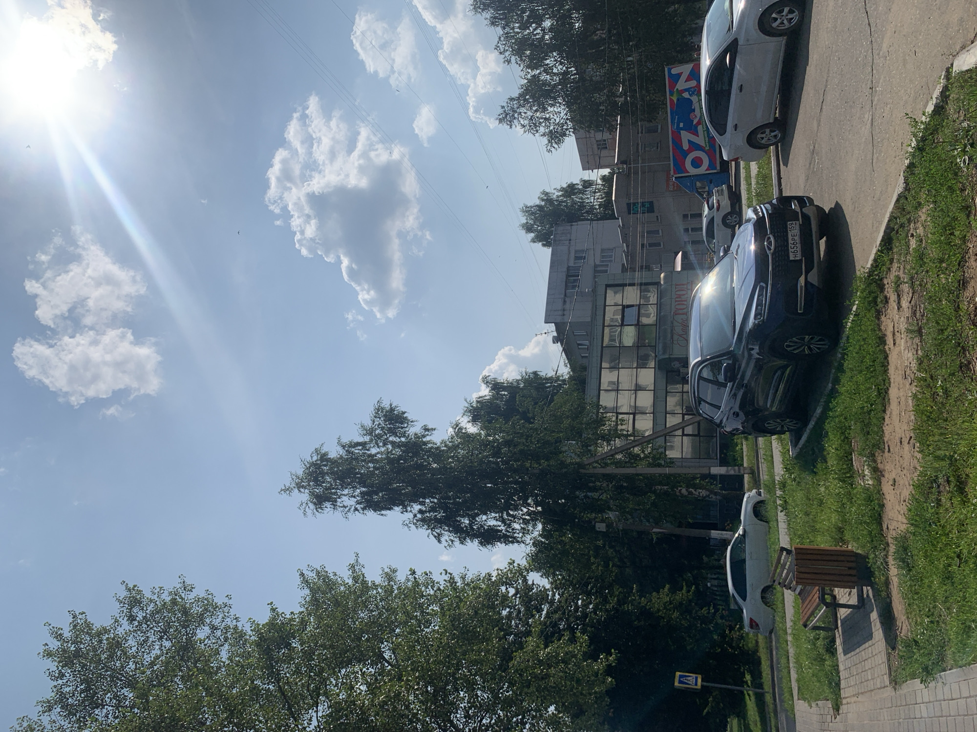 The image depicts a sunny day in Chaikovsky, Russia, with a clear blue sky punctuated by white, puffy clouds and the bright glare of the sun. The scene captures a street view with parked cars and buildings. In the foreground, a dark-colored SUV is parked on the right side of the road, with its license plate reading "H656PE 159". To its left, a white sedan is parked on the opposite side of the street. Further down the street, another white sedan is visible.
A sidewalk runs along the left side of the image, featuring a wooden bench and a trash bin. Adjacent to the sidewalk, a pedestrian crossing sign is visible. Behind the sidewalk, a grassy area is present, bordered by trees.
In the background, several multi-story buildings are visible. One building, with a glass facade, appears to be a commercial establishment, possibly with signage that includes the words "Кафе ТОПОЛ" (Cafe Topol). Another building features the prominent blue and white logo of "OZON". Trees line the street and surround the buildings, indicating a moderately urbanized environment with green spaces. The presence of overhead wires suggests an active street infrastructure. No people are clearly visible in the image.