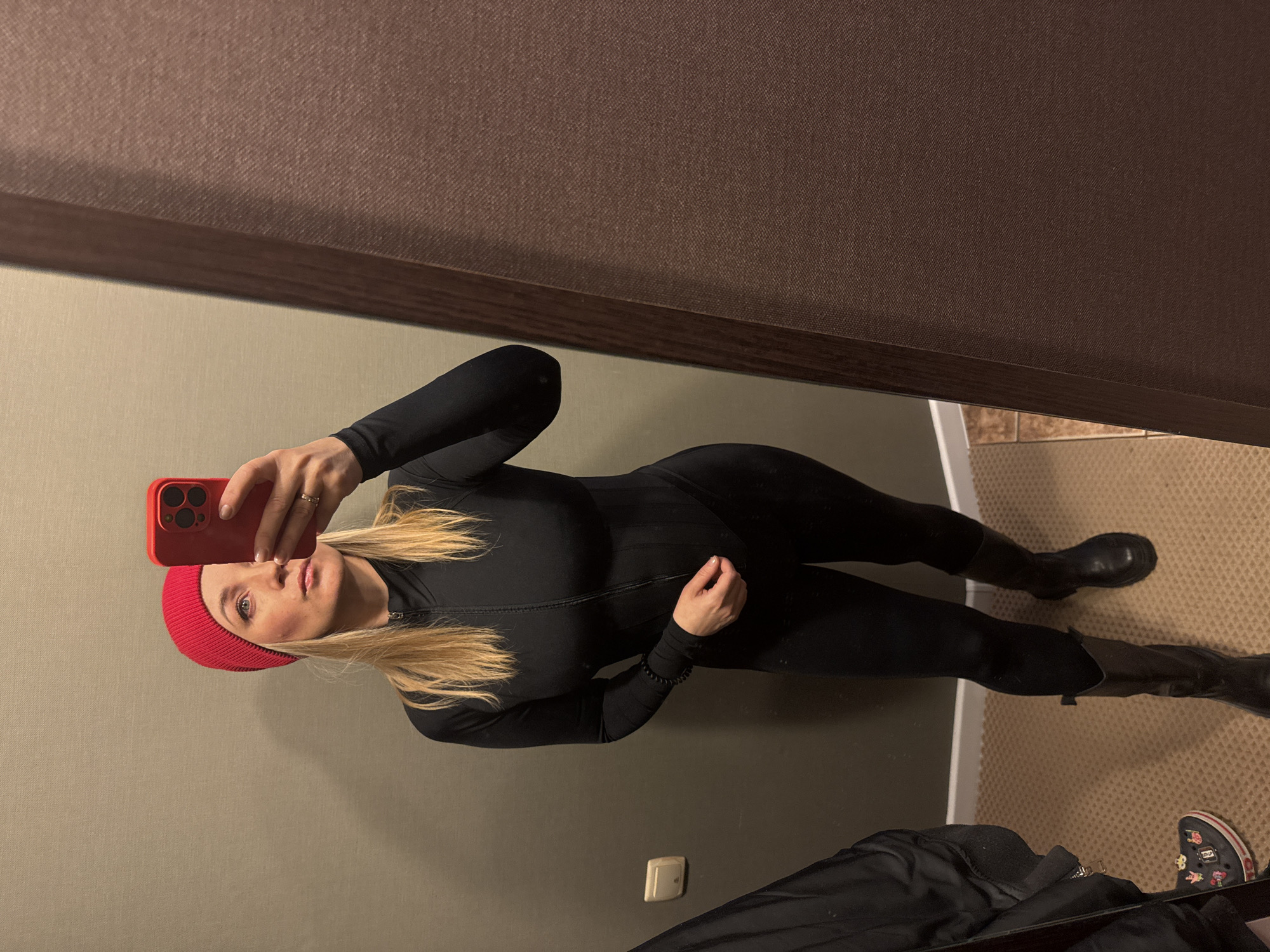 A woman wearing a red beanie, a black catsuit-like garment, and black boots stands in a room and takes a selfie in a mirror. The room has beige walls and carpet with a brown border near the floor. A black jacket and a pair of dark-colored shoes with cartoon decorations are visible on the floor. A brown frame surrounds the mirror. There is no information about the location in the city of Votkinsk, Russia.