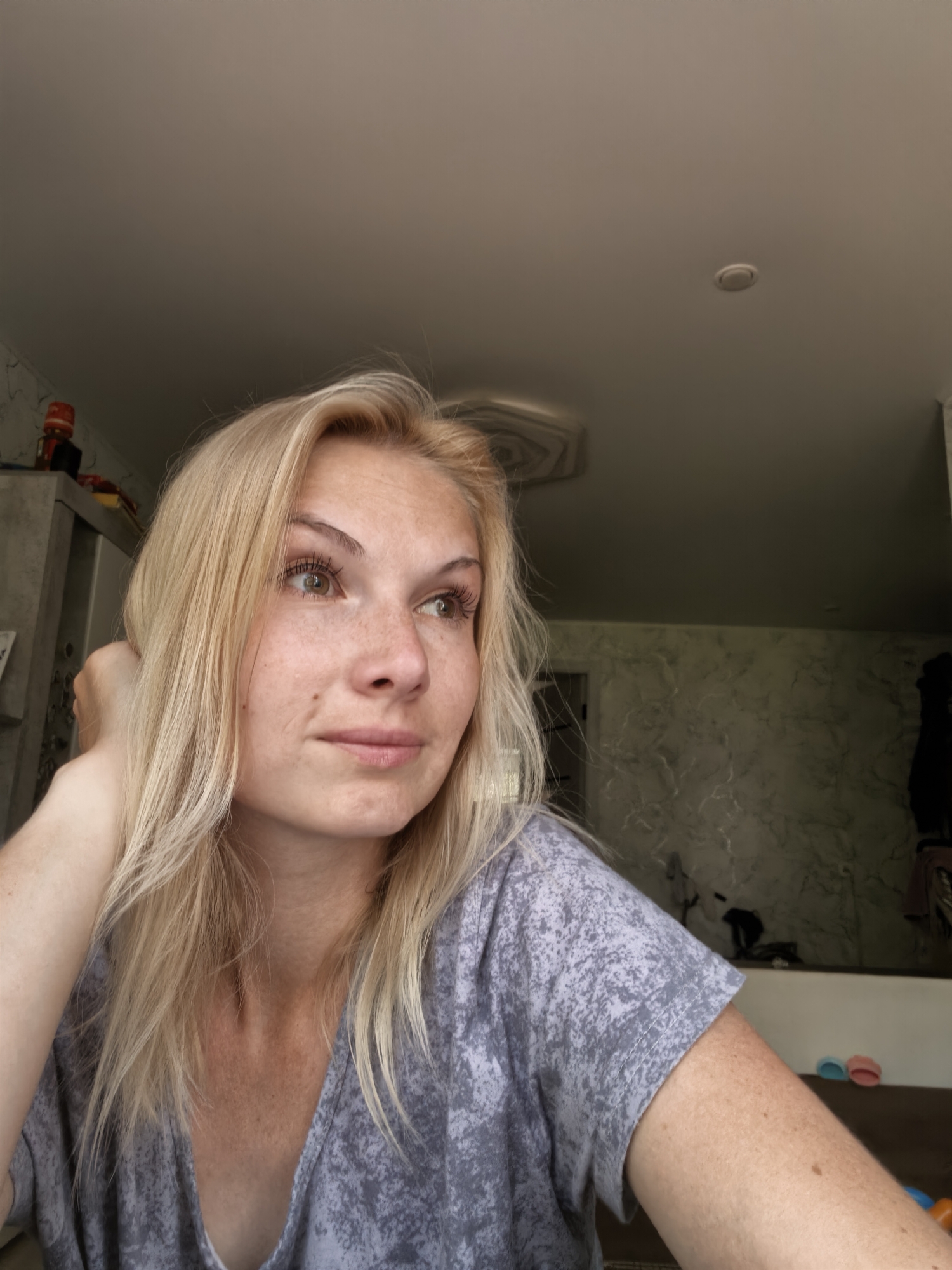 A young woman with blonde hair and freckles is looking to her right, with her left hand supporting her head. She is wearing a gray t-shirt with a speckled pattern. The background shows a ceiling with a light fixture, a wall with a marbled pattern, and some objects on shelves to the left. The setting appears to be indoors, possibly a residential interior in Sterlitamak, Russia.