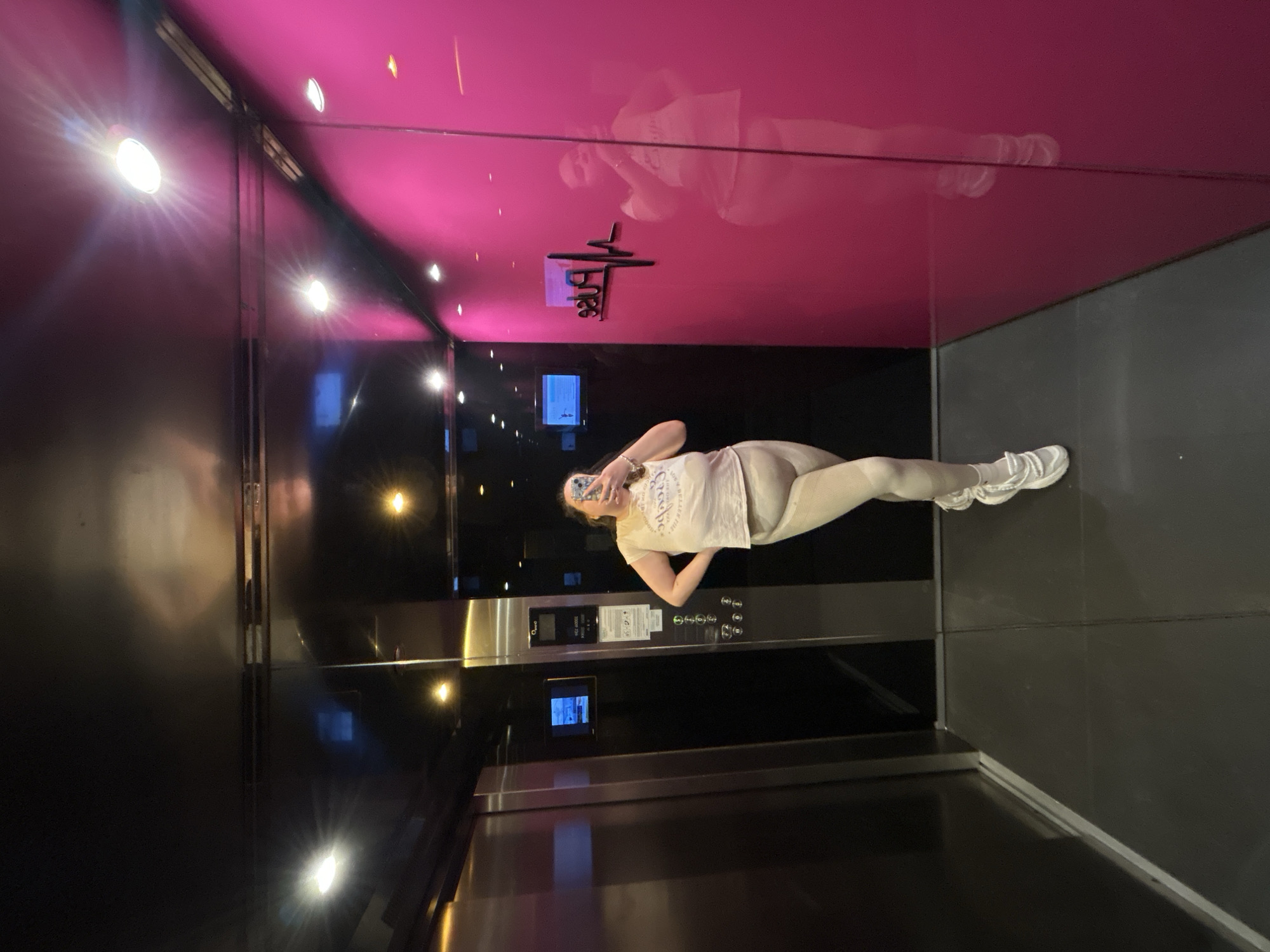 A young woman is taking a selfie in an elevator with a pink ceiling. She is wearing light-colored athletic clothing and white sneakers. The elevator has reflective walls and a control panel. The city of Plovdiv, Bulgaria is not discernible from the image.
