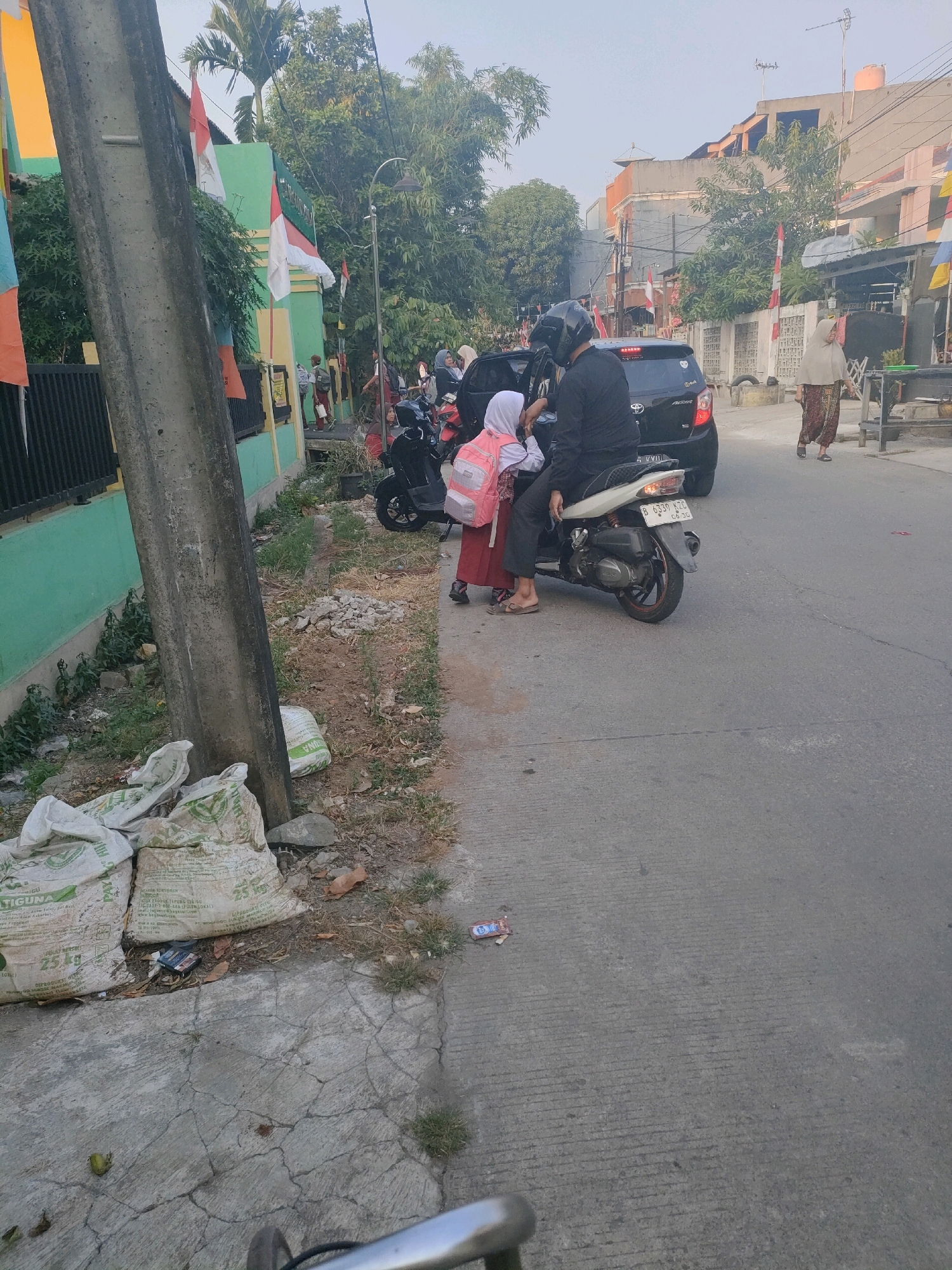 In Bekasi, Indonesia, on a street lined with buildings and trees, a man wearing a helmet sits on a motorcycle, with a young girl in a school uniform and backpack behind him. The girl is wearing a white hijab and a red skirt. They are parked next to a black car, and a woman in a headscarf walks in the background. On the left side of the image, a concrete pole stands next to a green building adorned with Indonesian flags. Several sacks of what appear to be fertilizer are piled on the ground near the pole. The street surface is asphalt, and there is debris scattered on the side of the road. The scene suggests a typical urban environment in Indonesia, possibly during the morning or late afternoon, given the lighting.