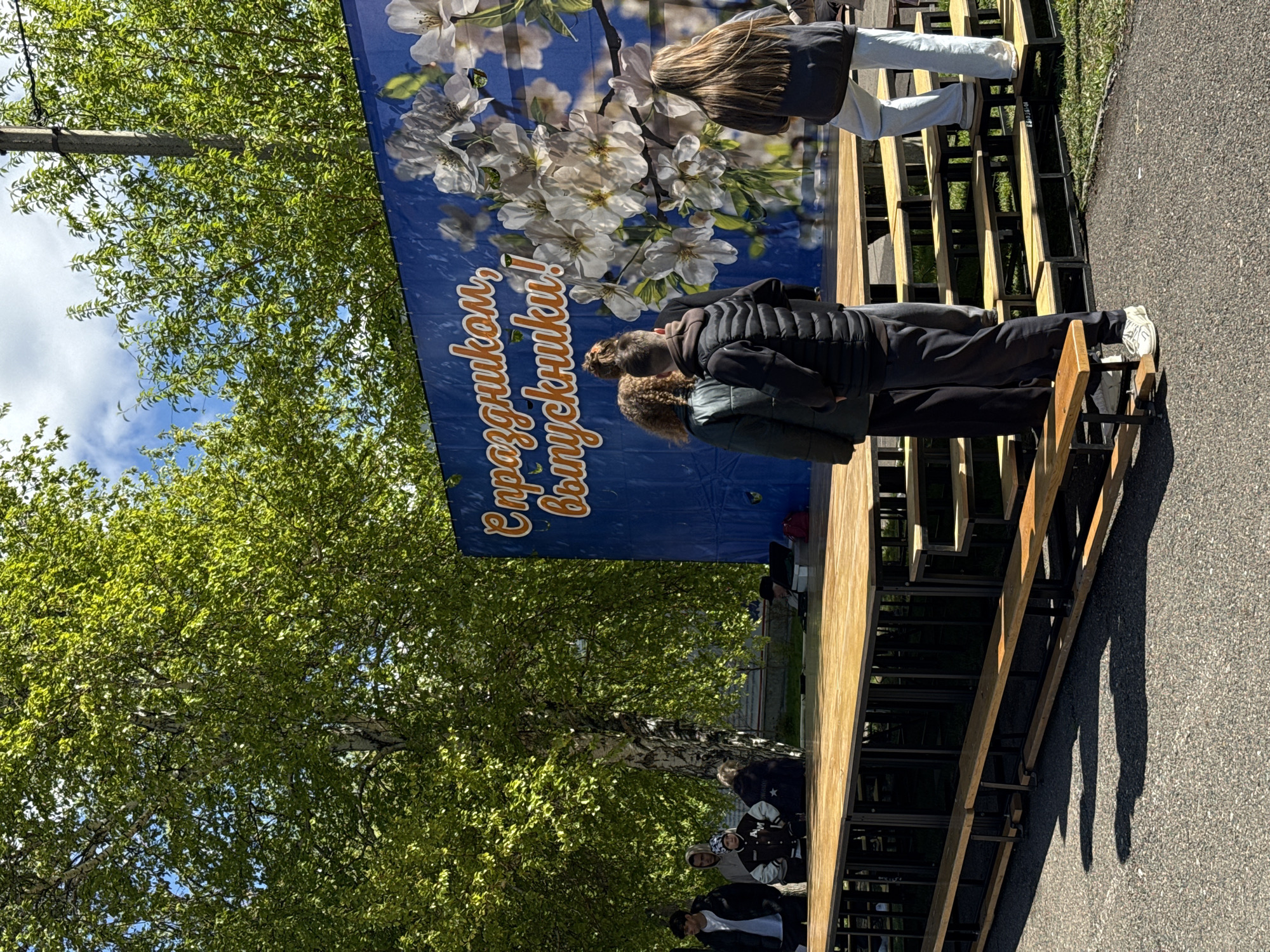 The image shows a temporary stage or viewing platform set up outdoors, likely for an event. The location appears to be Krasnoyarsk, Russia, judging by the Cyrillic text on the banner ("С праздником, выпускники!") which translates to "Happy graduation, graduates!".
**People:** Several people are present. Two are standing on the platform. A group of people are standing on the ground level.
**Objects:** A stage is constructed with wooden planks. A banner with text and floral images hangs behind the platform. Trees are visible in the background.
**Environment:** The setting is outdoors, with trees, suggesting a park or urban green space. The ground appears to be paved. The time of day appears to be daytime.