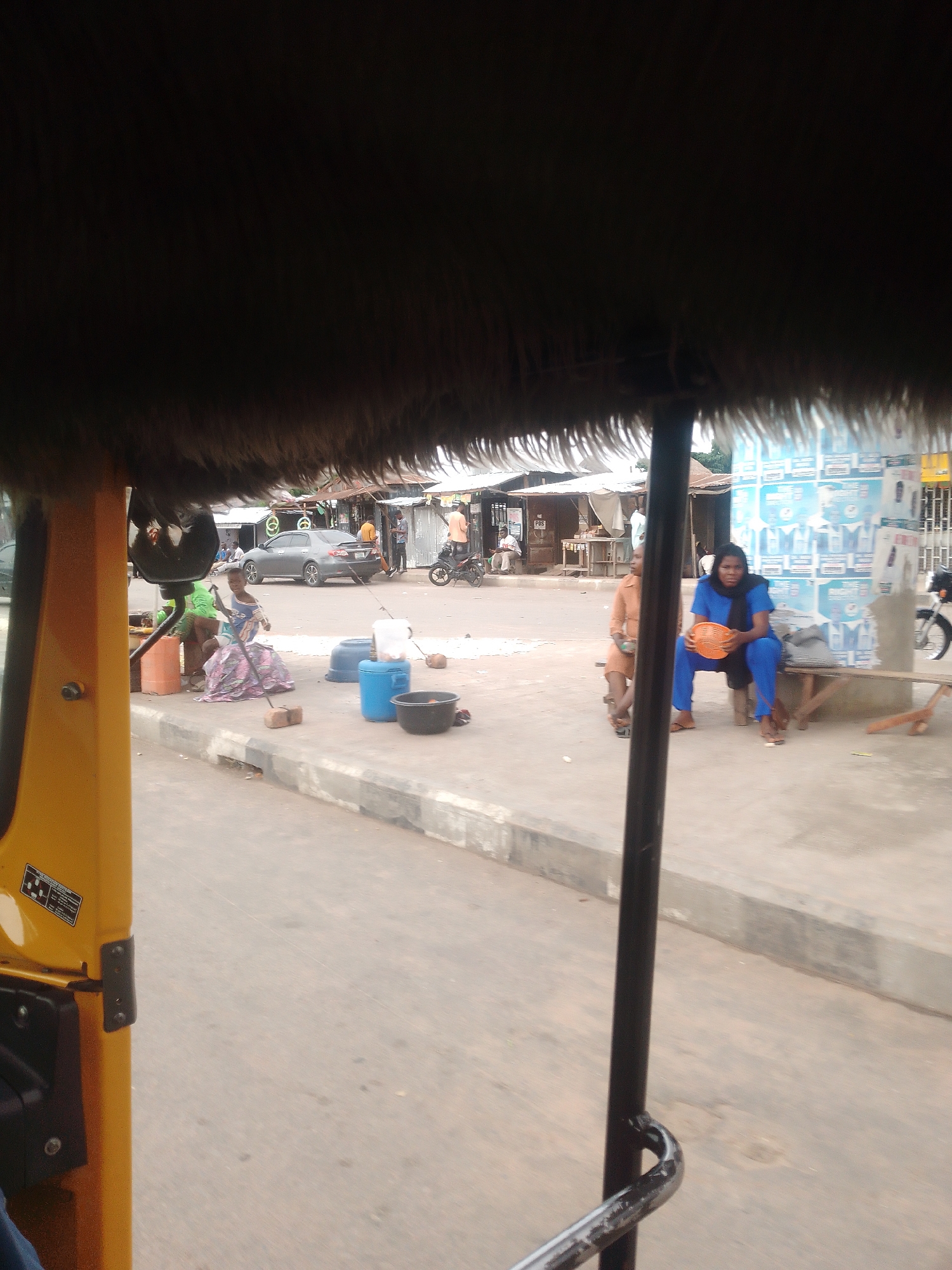 The image depicts an outdoor scene in what appears to be a market or roadside area. In the foreground, the edge of a yellow vehicle, likely an auto-rickshaw, is visible, with a furry trim along its roof.
Several people are present. To the left, a woman in a green top and patterned skirt is seated with several orange containers around her. Next to her, another person, also seated, is partially obscured. Further to the right, a woman in an orange dress is sitting on a wooden bench. Beside her, a woman in a blue outfit is holding an orange basket. Behind them, a gray car is parked, and several other individuals are standing and interacting in the background. A motorcycle is also parked on the right side of the frame.
The setting includes a paved road and a sidewalk. Various containers, including large blue and black buckets, are scattered on the ground. Buildings with corrugated metal roofs are visible in the background, suggesting a commercial or residential area. Several posters or advertisements are displayed on a wall behind the people on the bench.
The time of day appears to be daytime, and the weather seems overcast or hazy. The overall impression is of a bustling, everyday scene in a Nigerian town. There is no discernible text of significance within the visible portion of the image.