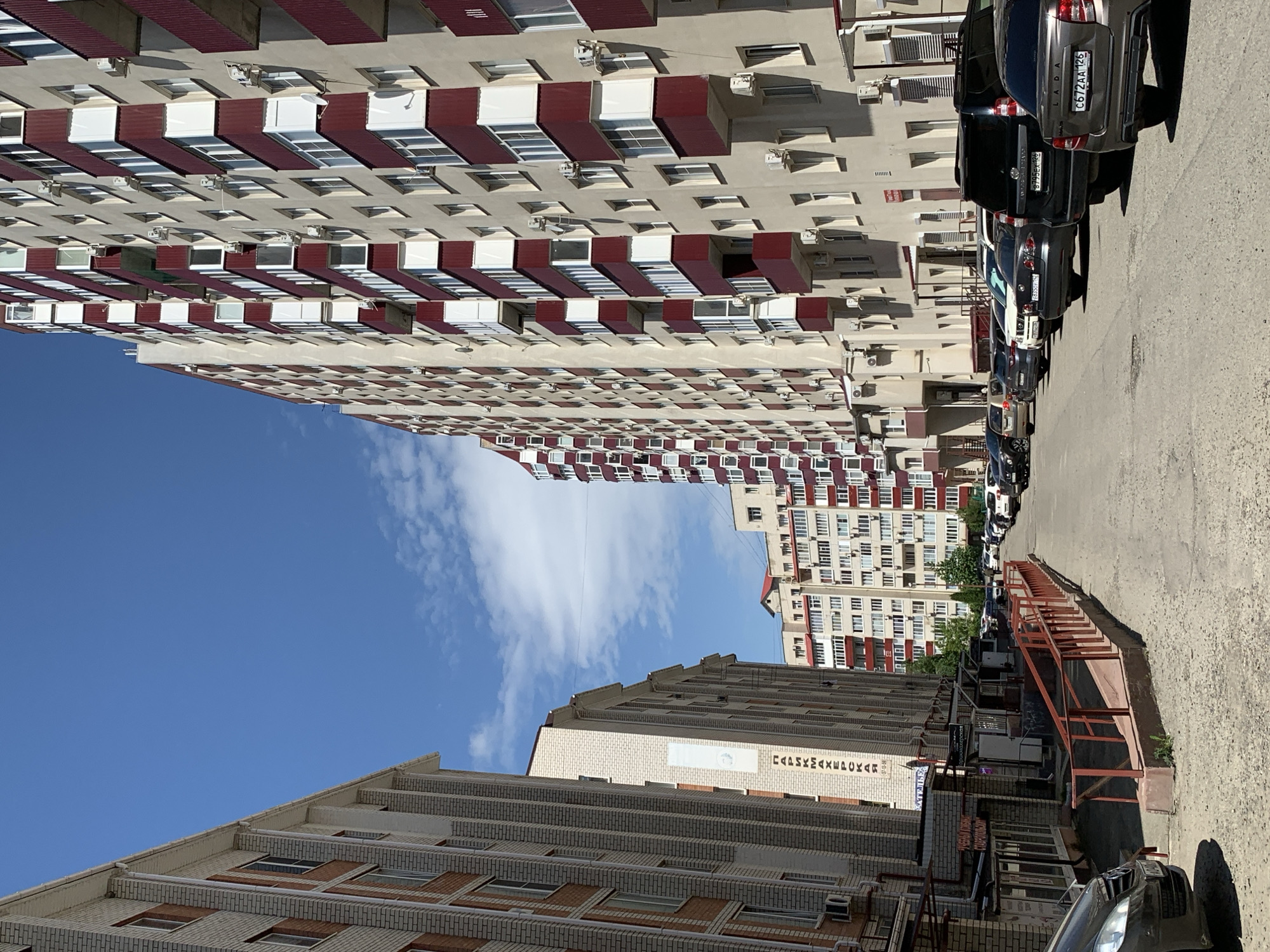 The image depicts a street scene in Stavropol, Russia, showcasing a mix of residential and commercial buildings. Two tall apartment buildings dominate the view, one with a repeating pattern of white windows and red balconies, and the other with brick and light-colored facades. The sky is clear blue with some clouds. A row of parked cars lines the right side of the street, while commercial storefronts with a red railing are on the left.