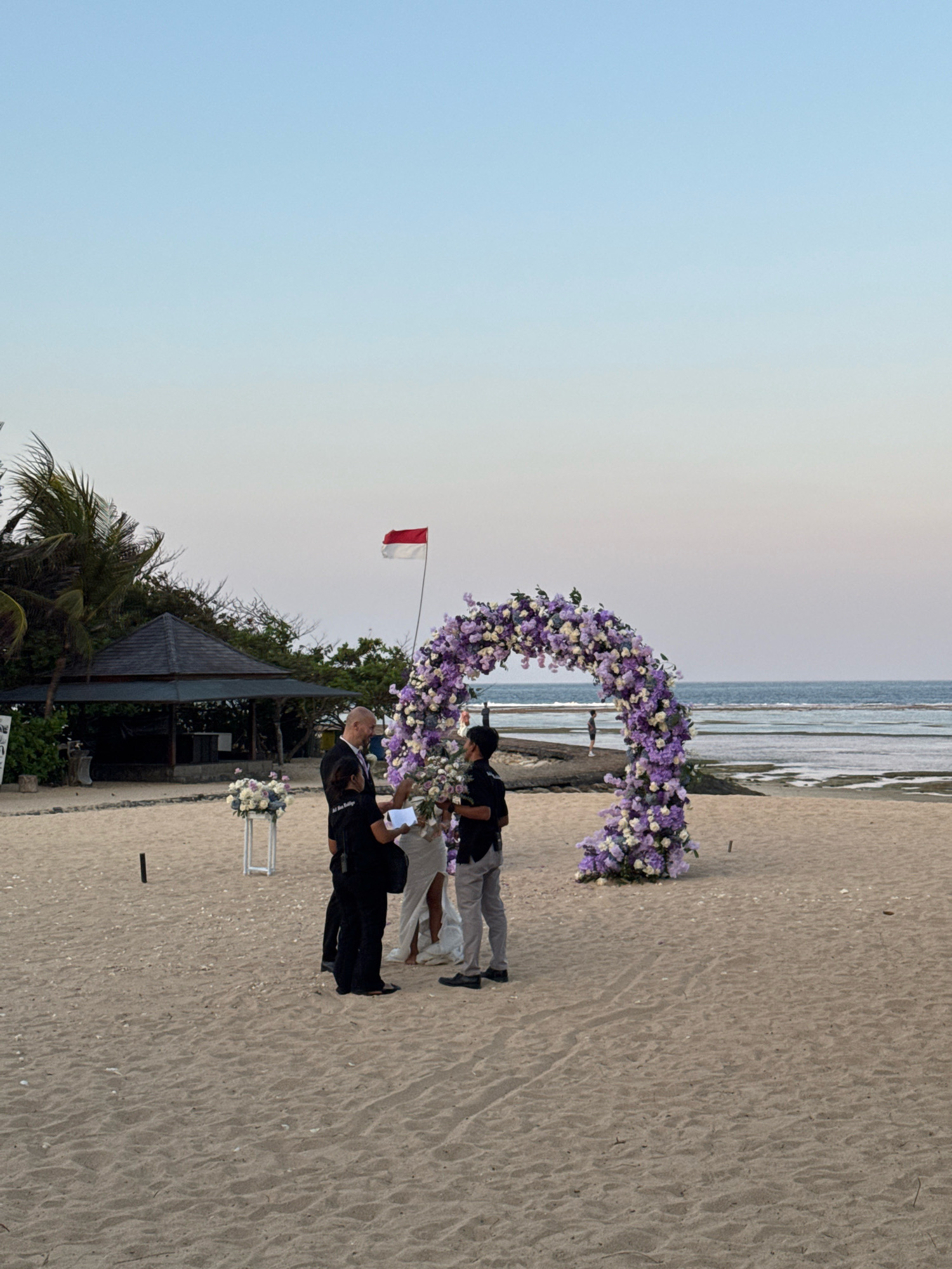 An outdoor wedding ceremony is taking place on a sandy beach in Kuta Selatan, Indonesia, with the ocean visible in the background under a clear, pale blue sky. In the foreground, a floral archway, adorned with purple and white flowers, stands as a centerpiece for the ceremony. Several individuals are gathered near the archway. A man in a dark suit appears to be officiating the ceremony, facing a couple. The bride, in a white dress, and the groom are positioned in front of him. Two other men in dark clothing are also present, possibly assisting with the ceremony or arrangements. A red and white Indonesian flag flutters on a pole to the left of the archway. To the far left, a small thatched-roof structure is visible, surrounded by palm trees and greenery. White flower arrangements are placed on stands in front of the structure. The sand is lightly textured with visible footprints and tire tracks. The tide is out, revealing rocky formations in the shallow water.