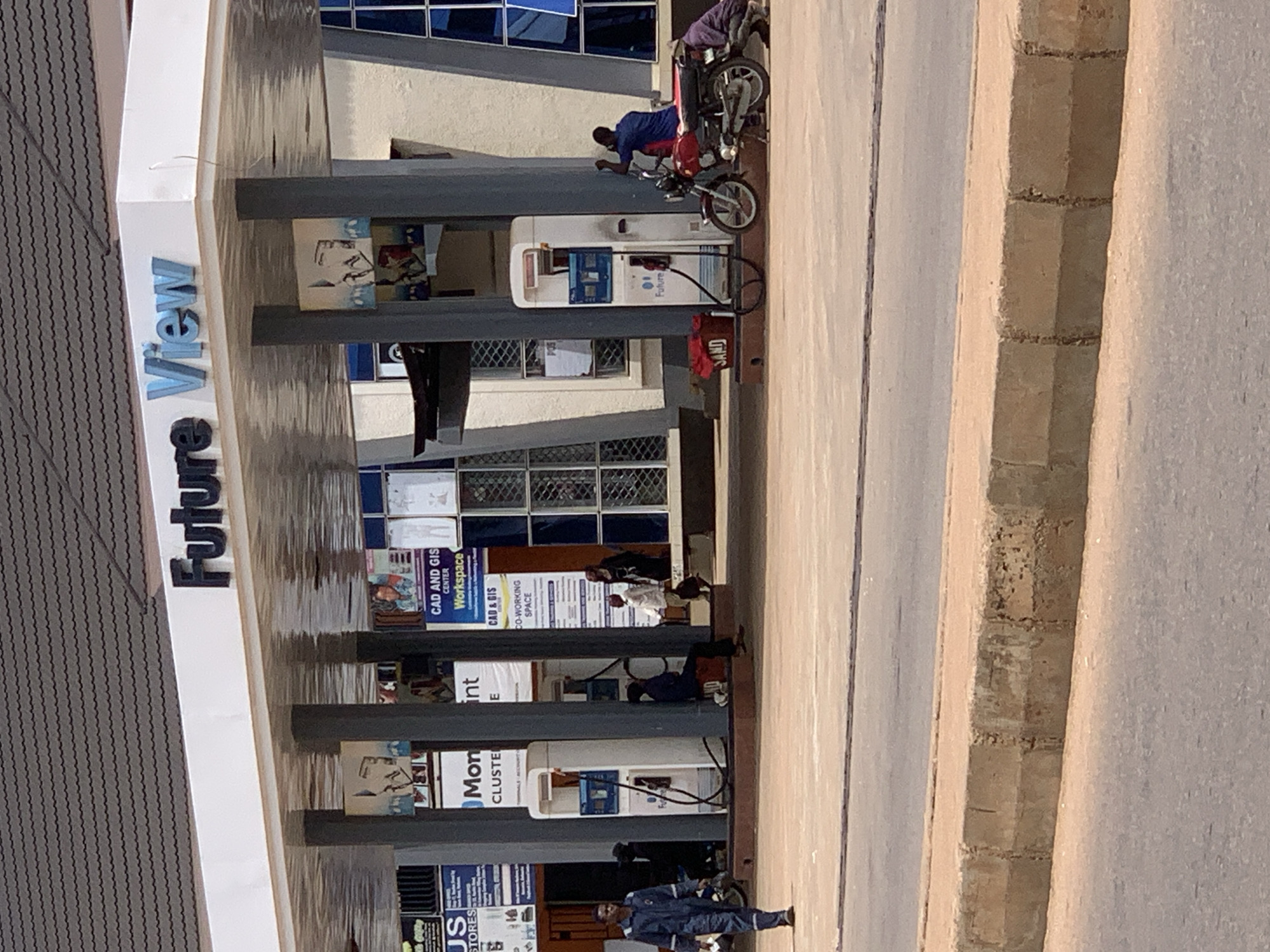 The image shows a street-level view of a "Future View" gas station in Sabon Tasha, Nigeria. The gas station has a white canopy supported by grey pillars. There are several gas pumps visible, some with hoses attached. On the left side of the image, a man in a blue uniform is walking away from the camera. In front of one of the gas pumps, a red bucket labeled "SAND" is present. A red motorcycle is parked to the right of another gas pump, with two individuals interacting around it; one is sitting on the motorcycle, and the other is kneeling beside it. The background shows the facade of a building with windows, some of which are tinted blue. The ground in front of the gas station appears to be paved, with a curb and road in the foreground. The overall scene depicts a typical day at a gas station in a Nigerian city.