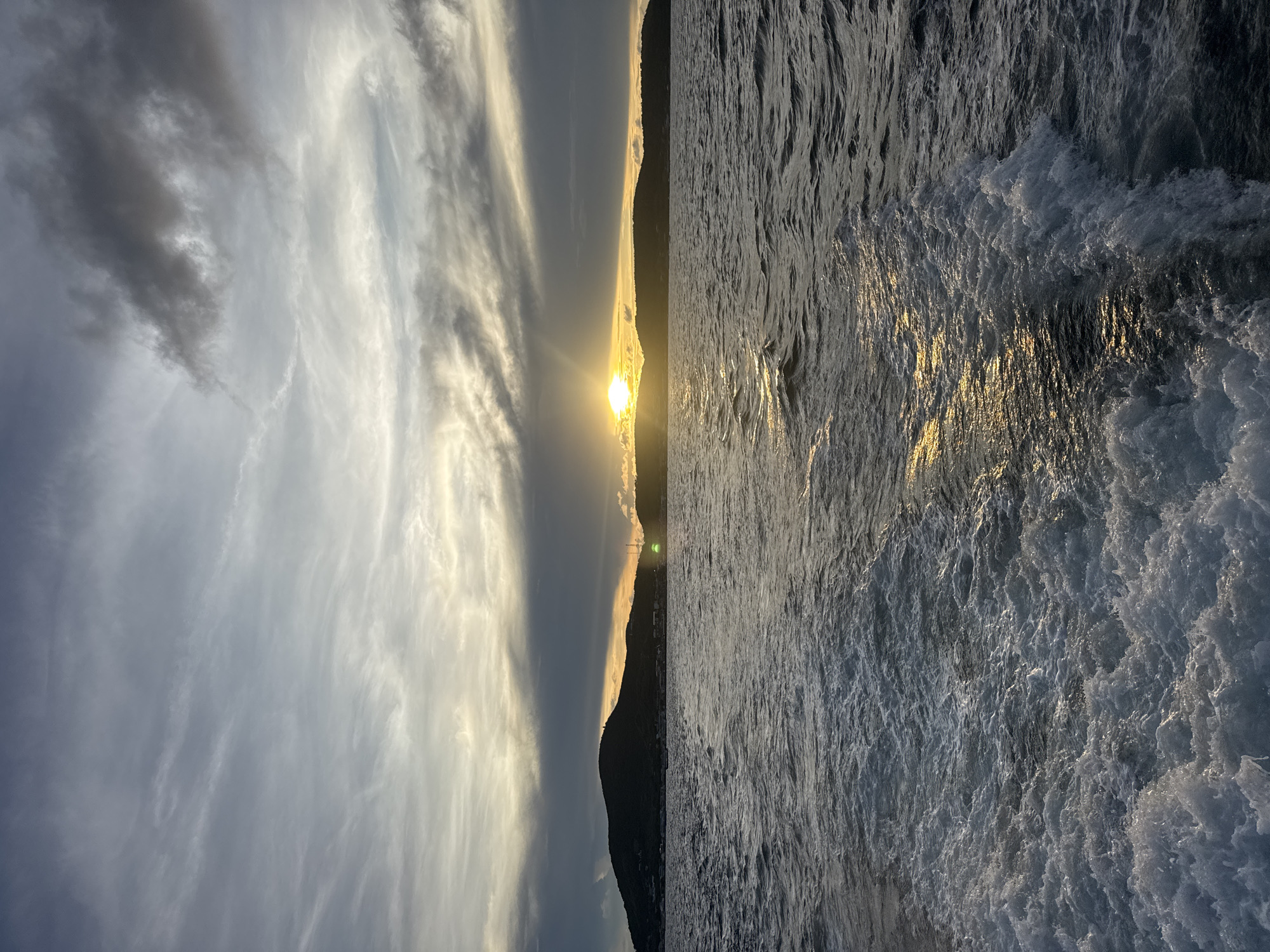 The image shows a seascape at sunset in Pattaya City, Thailand. A low-lying coastline silhouette is visible, separating the sky and the ocean. The sun is positioned at the horizon line, its glow diffused by thin cloud layers. The sky displays a mix of light and dark clouds, ranging from light grays to darker hues. The ocean surface is textured with waves, and the sunlight reflects off the water, creating a bright, shimmering effect. There are no discernible people or objects beyond the natural elements. The interaction is between light and water, creating a scenic vista.