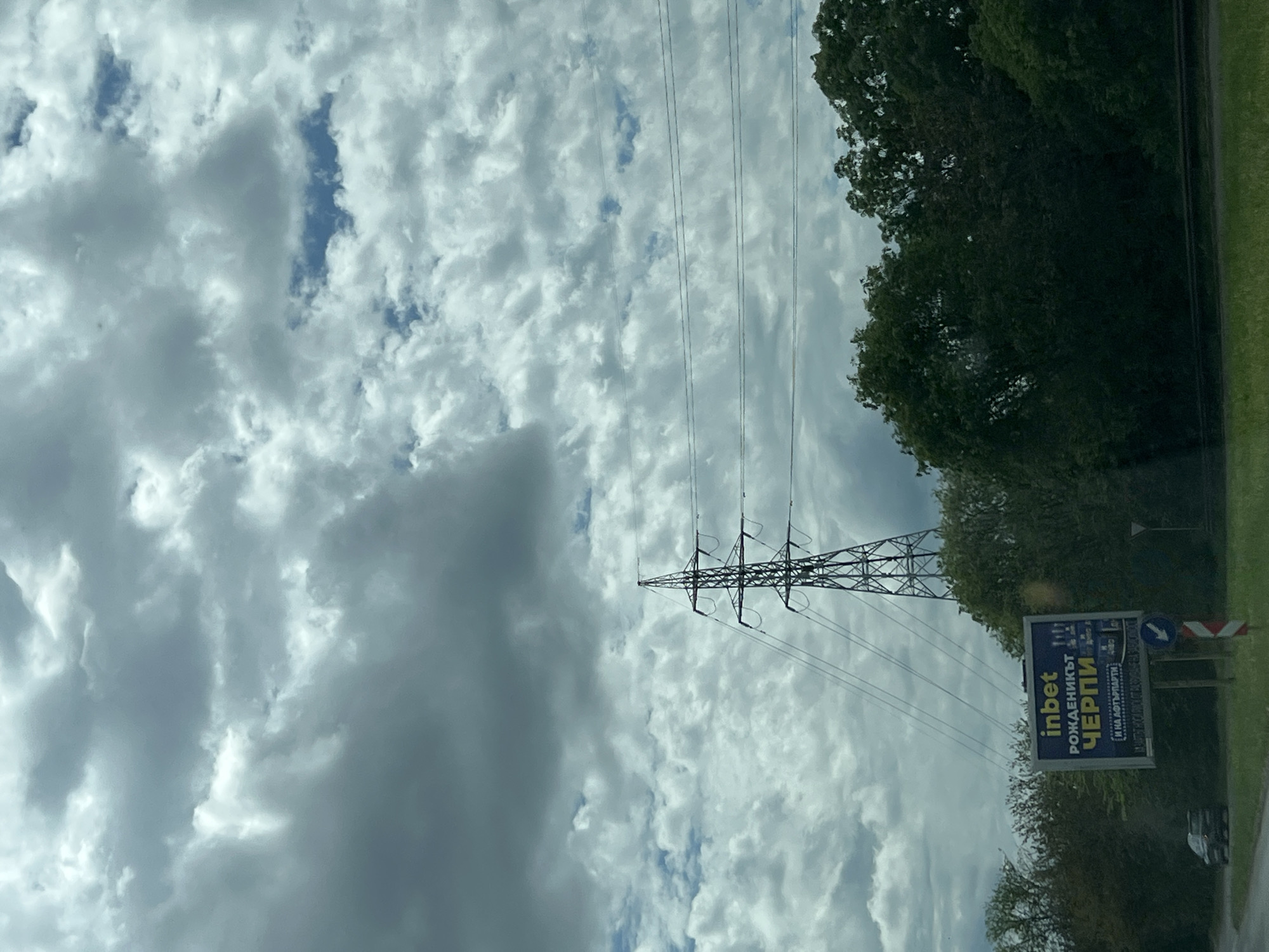 The image shows a roadside scene with a cloudy sky in Burgas, Bulgaria. An electrical transmission tower and power lines dominate the center of the frame. To the right is a sign with the word "inbet" and other Bulgarian text, partially obscured by trees and vegetation. A road and car are barely visible in the lower right corner. The overall environment suggests an area on the outskirts of the city, possibly near a highway or major road.