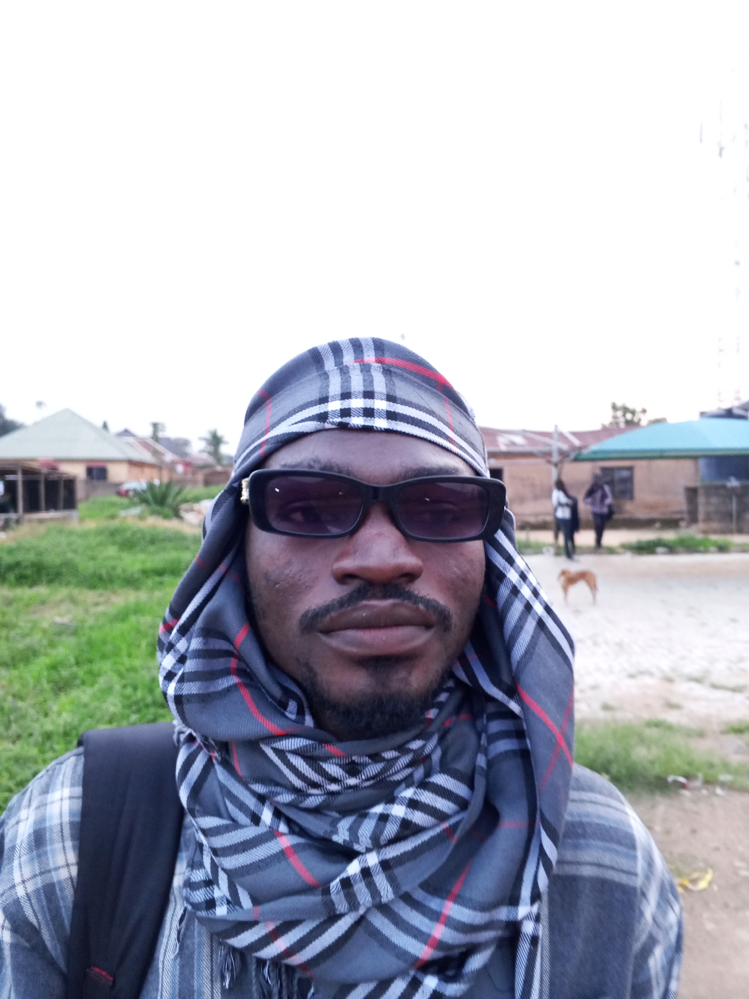 A man wearing a plaid scarf and sunglasses is pictured in Bukuru, Nigeria. He has a beard and is looking at the camera. He is wearing a plaid shirt and a backpack. In the background, there are buildings and greenery. A dog is visible in the distance. The sky is overcast.