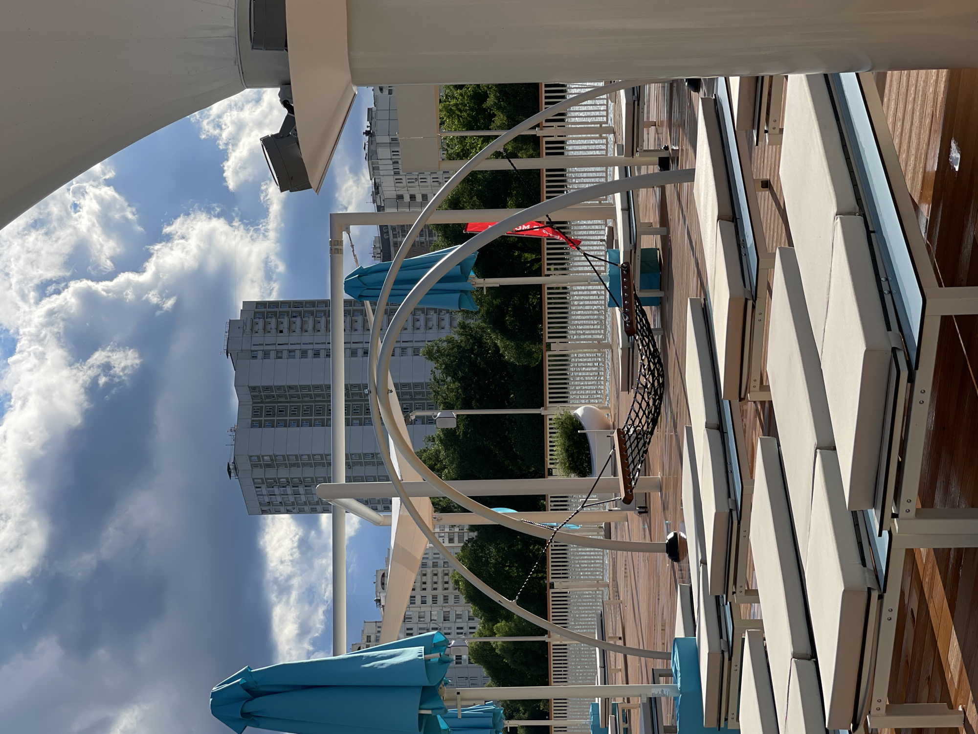 This image depicts an outdoor area, possibly a rooftop or deck, in Moscow, Russia. The scene is set on a sunny day with scattered clouds. In the foreground, there are several white lounge chairs with beige cushions arranged on a wooden deck. A large white structure, possibly a sail or awning, arches over the scene, casting shadows.
Behind the lounge chairs, a white railing separates the deck from a background of lush green trees. Beyond the trees, several tall, modern white buildings are visible, characteristic of urban architecture. The sky is a bright blue, interspersed with fluffy white and darker grey clouds, suggesting dynamic weather.
Two bright turquoise umbrellas are partially visible in the lower left corner of the image, suggesting the area is intended for relaxation and perhaps shade from the sun. A black net hammock hangs from a metal frame, adding another element of leisure. A small red flag with some white lettering is also visible near the hammock, though the text is not clearly legible.
The overall impression is one of a modern, well-equipped outdoor space designed for comfort and enjoyment, set against a backdrop of a bustling city. The lighting and cloud formations suggest it's daytime, likely late morning or afternoon.