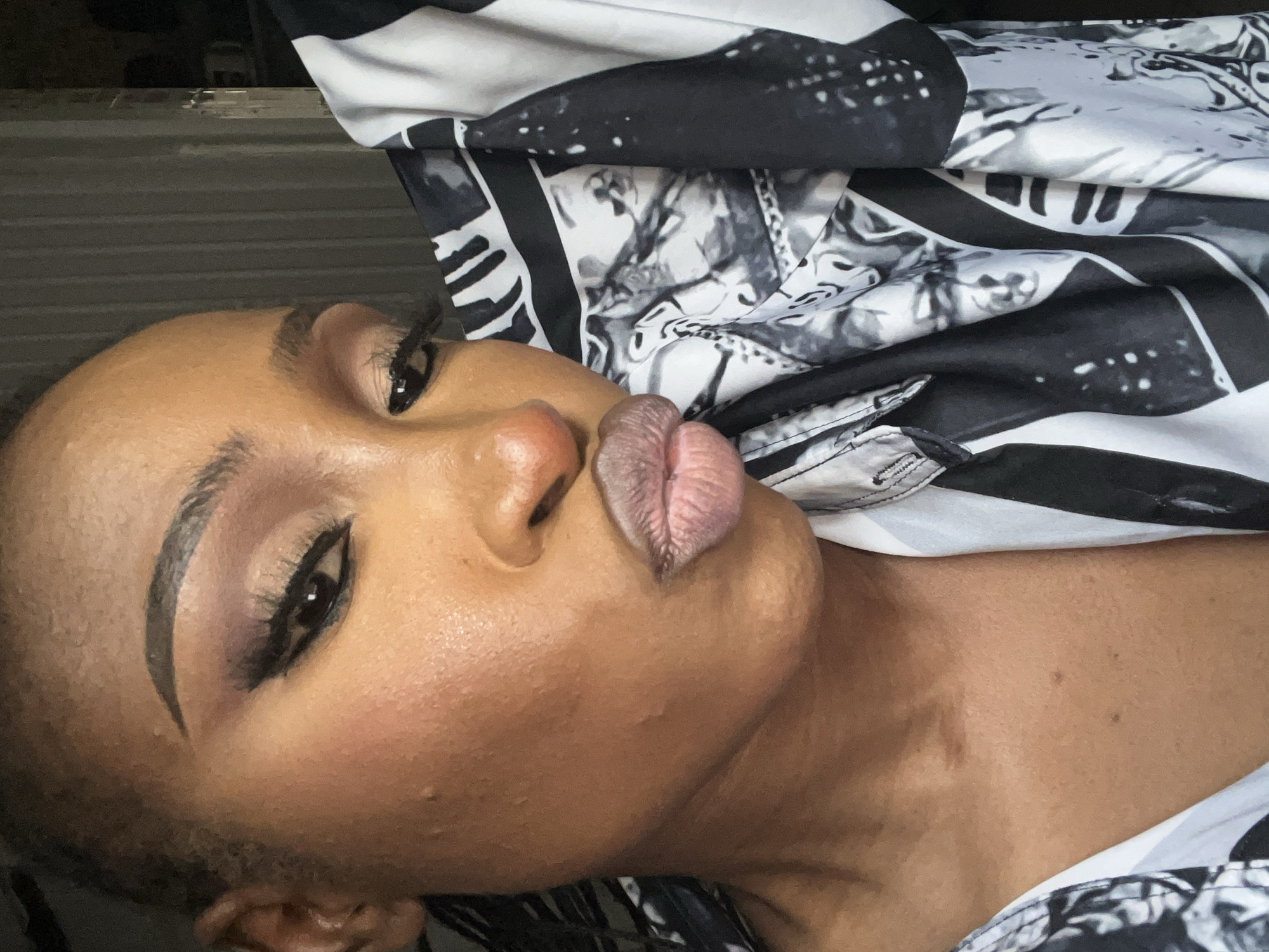 A close-up selfie shows a woman of color wearing makeup and a black and white patterned shirt. Her face is turned to the side, and her lips are pursed. The background is dark and out of focus, suggesting an indoor setting, possibly a studio or a room in a residential or commercial building in Abuja, Nigeria. The lighting is soft, highlighting her features and the texture of her skin and the fabric of her shirt. The overall impression is one of a casual and confident self-portrait.