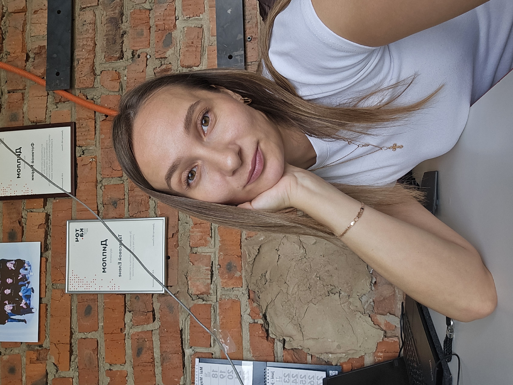 A woman with light brown hair is leaning her head on her hand in a photograph. She is wearing a white t-shirt and a gold necklace with a cross pendant. A gold bracelet is visible on her wrist. The background features a brick wall with two framed certificates and a picture. The certificate on the left has the word "Моппид" written on it, and the one on the right has "NNGE NOHΝΤΟΦ." A calendar is on the desk in front of the woman. The location is Chaikovsky, Russia.