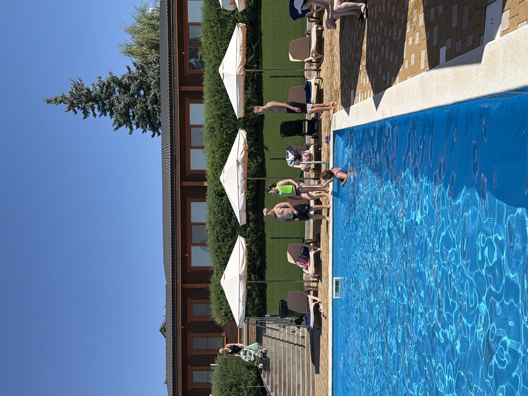 The image depicts a sunny day at an outdoor swimming pool area. A large blue swimming pool with shimmering water occupies the foreground and mid-ground. The pool is bordered by a light-colored concrete edge and a patio paved with light and dark brown bricks. Several lounge chairs are placed around the pool, some with towels and personal items on them. Two large white umbrellas provide shade.
In the background, a long, dark wooden building with multiple windows is visible. A dense green hedge runs along the front of the building, separating it from the pool area. A tall evergreen tree stands behind the building.
On the left side of the image, a woman in a black swimsuit is pushing a stroller up a wide set of concrete stairs. Several people are standing and sitting in the area around the pool. One man, wearing black shorts, stands near the pool's edge. A young boy in black shorts is also visible. A woman in a blue swimsuit is partially submerged in the pool. Two women are sitting on a bench on the right side of the image, with one wearing a white cap.
The image appears to have been taken in the city of Kamyshin, Russia, likely at a resort or recreational facility, given the presence of the pool, lounge chairs, and the building in the background. The overall scene suggests a leisurely summer day.