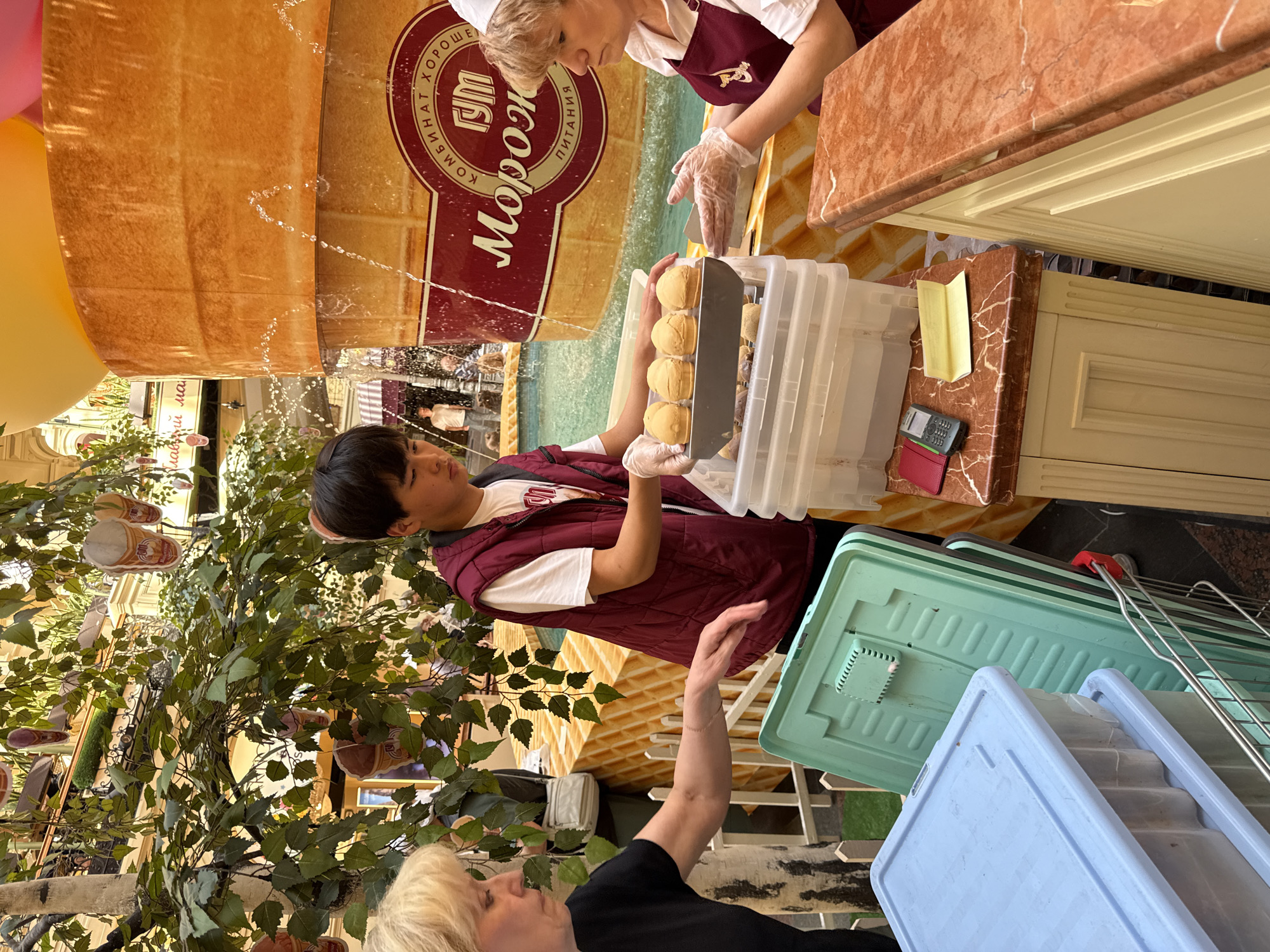 The image depicts an indoor scene at what appears to be a food stall or concession stand, likely within a large public building such as a mall or grand hall, indicated by visible escalators and decorative elements in the background. The primary activity centers around a transaction involving three individuals.
Dominating the background is a large, textured facade designed to resemble a giant waffle cone. Decorative water droplets cascade down its surface, suggesting a fountain feature. A prominent circular red and white logo is affixed to this facade, displaying Cyrillic text that reads "КОМБИНАТ ХОРОШЕЕ МОРОЖК ПИТАНИЯ," which roughly translates to "GOOD FOOD COMBINE ICE CREAM NUTRITION" or similar, strongly suggesting an ice cream or dessert vendor.
Behind a light-brown marble-patterned counter, a woman with blonde hair, wearing a white cap or bandana, a maroon apron, and clear gloves, leans forward, seemingly in the process of serving or supervising. Facing her is a young man with dark hair, dressed in a white t-shirt and a maroon vest, also wearing clear gloves. He holds a metal tray containing several light-colored, round-shaped baked goods, possibly small waffles or pastries. On the counter, beside clear plastic containers holding more of these items, is a small black and red cash register or calculator.
In the immediate foreground, a second blonde-haired woman, wearing a black top, is partially visible, extending her arm towards the scene. Green artificial plants and trees are strategically placed, enhancing the indoor environment. On the floor, light blue and teal plastic crates are stacked. Small decorative ice cream cone shapes are suspended from the ceiling in the background. The scene is brightly lit, indicative of daytime. Based on the Cyrillic text and context, the location is Moscow, Russia.
