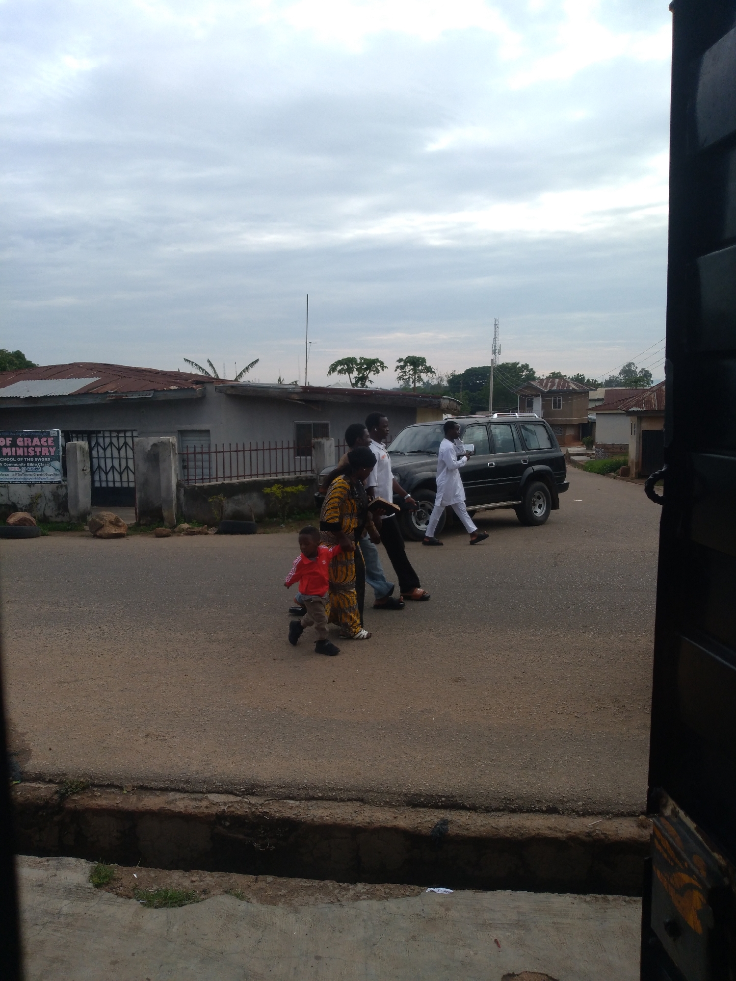 The image depicts a street scene under an overcast sky in a residential or semi-urban area within the city of Jos, Nigeria. The lighting is diffused and uniform. In the immediate foreground, an open concrete drainage ditch is visible, with sparse vegetation growing within it. The viewpoint is from a slightly elevated position, possibly an open doorway or gate, indicated by the dark, partially visible frame on the far right. The main subject is a group of at least five individuals walking on an asphalt road that appears to curve slightly to the right. From left to right: 1. A young child, wearing a red long-sleeved shirt, light-colored trousers, and dark shoes, is in the foreground, taking a step forward with their left leg, looking towards the left side of the frame. 2. Immediately behind the child, a woman wearing a yellow and brown patterned dress and light-colored sandals is walking. Her left arm is extended towards the child, suggesting she is accompanying or guiding them. 3. To the right and slightly behind the woman, a man wearing a white t-shirt, dark trousers, and dark sandals is walking with his left foot forward. He holds a dark object in his left hand. 4. Further to the right, a black Toyota Land Cruiser SUV, likely an 80 series, is stationary or moving slowly, facing towards the right side of the frame. It features a roof rack. 5. Walking alongside the SUV, a man dressed entirely in white traditional Nigerian attire (a long-sleeved shirt and trousers) and dark shoes is depicted mid-stride, moving towards the right. 6. A sixth individual, largely obscured by the left edge of the frame, is partially visible wearing a white shirt and blue jeans, walking towards the right. On the left side of the road, a low, single-story building with a rusty corrugated metal roof is visible. In front of this building, a white sign with blue text reads "OF GRACE MINISTRY" and "SCHOOL OF THE SWORD / A COMMUNITY BIBLE CLASSI". A fence with concrete pillars and dark metal railings secures the property, with a dark metal gate visible. Two dark vehicle tires are placed on the ground near the fence, along with several large rocks. In the background, on both sides of the road, various residential structures with corrugated metal roofs are visible. A tall, lattice-structure communications tower stands prominently in the mid-ground, behind the