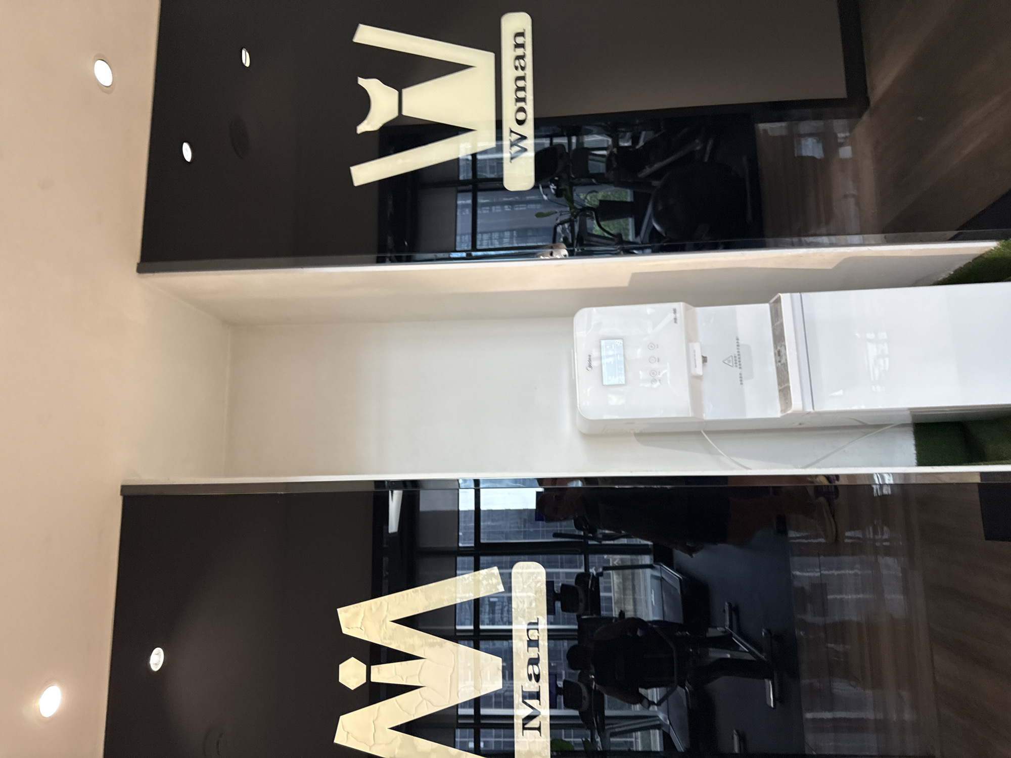 The image shows the entrance to men's and women's restrooms in a gym, likely located in Haizhu District, China. The doors are black glass with white stylized pictograms and text. The men's restroom door on the left features a stylized 'M' and the word "Man". The women's restroom door on the right features a stylized 'W' that resembles a dress, and the word "Woman". Inside the gym, treadmills and other exercise equipment are visible through the glass doors. A white water dispenser is positioned between the two doors. Reflections in the glass show people inside the gym and the photographer.