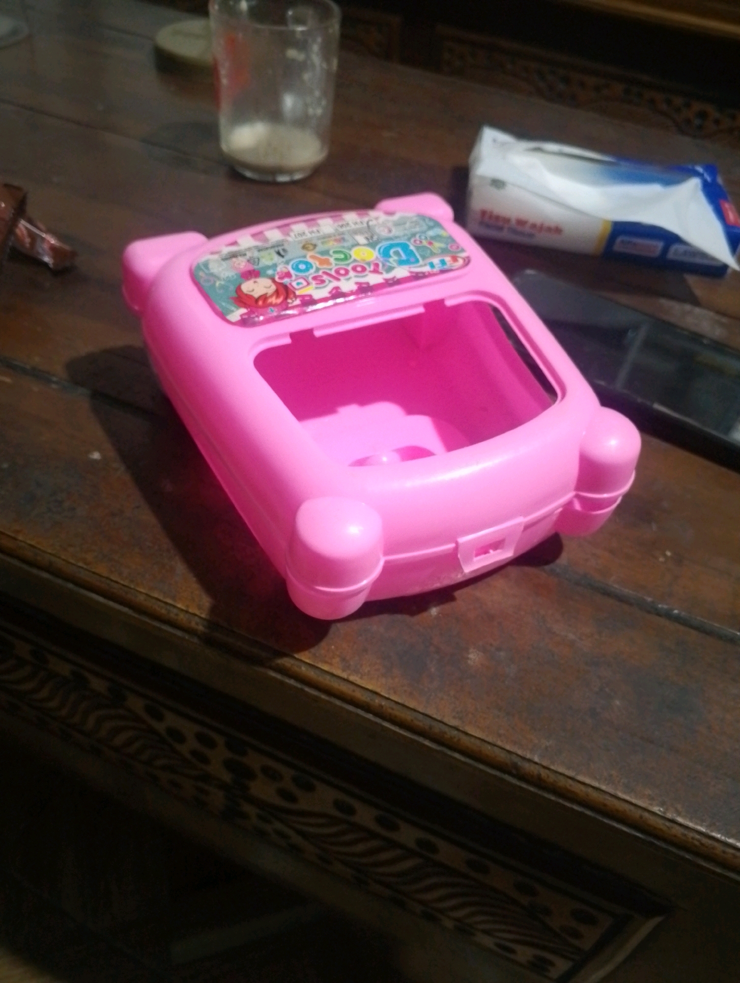 A pink toy appears to be the main subject of the photograph, positioned on a wooden table. The toy has a somewhat rectangular, open-top design with rounded edges and decorative stickers featuring cartoon characters and colorful text. To the left of the toy, a clear glass with some liquid and sediment is visible, as is a small piece of what looks like chocolate or candy. In the background, a package of tissues, identified by the text "Tisu Wajah," is partially obscured by the toy. A dark rectangular object, possibly a phone, is also present on the table behind the toy. The wooden table exhibits a textured surface with a decorative border underneath. The lighting suggests an indoor setting, with shadows cast by the objects. The image was captured in Kendalrejo, Indonesia.