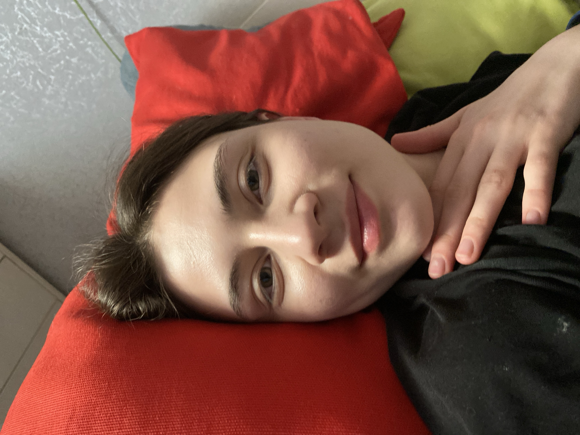 A young woman lies on her side, her head resting on a red pillow. She appears to be wearing a dark-colored garment. Her right hand rests on her chest, near her neck. The background includes a yellow pillow and a wall. There is no information available to confirm a location in the city of Votkinsk, Russia.