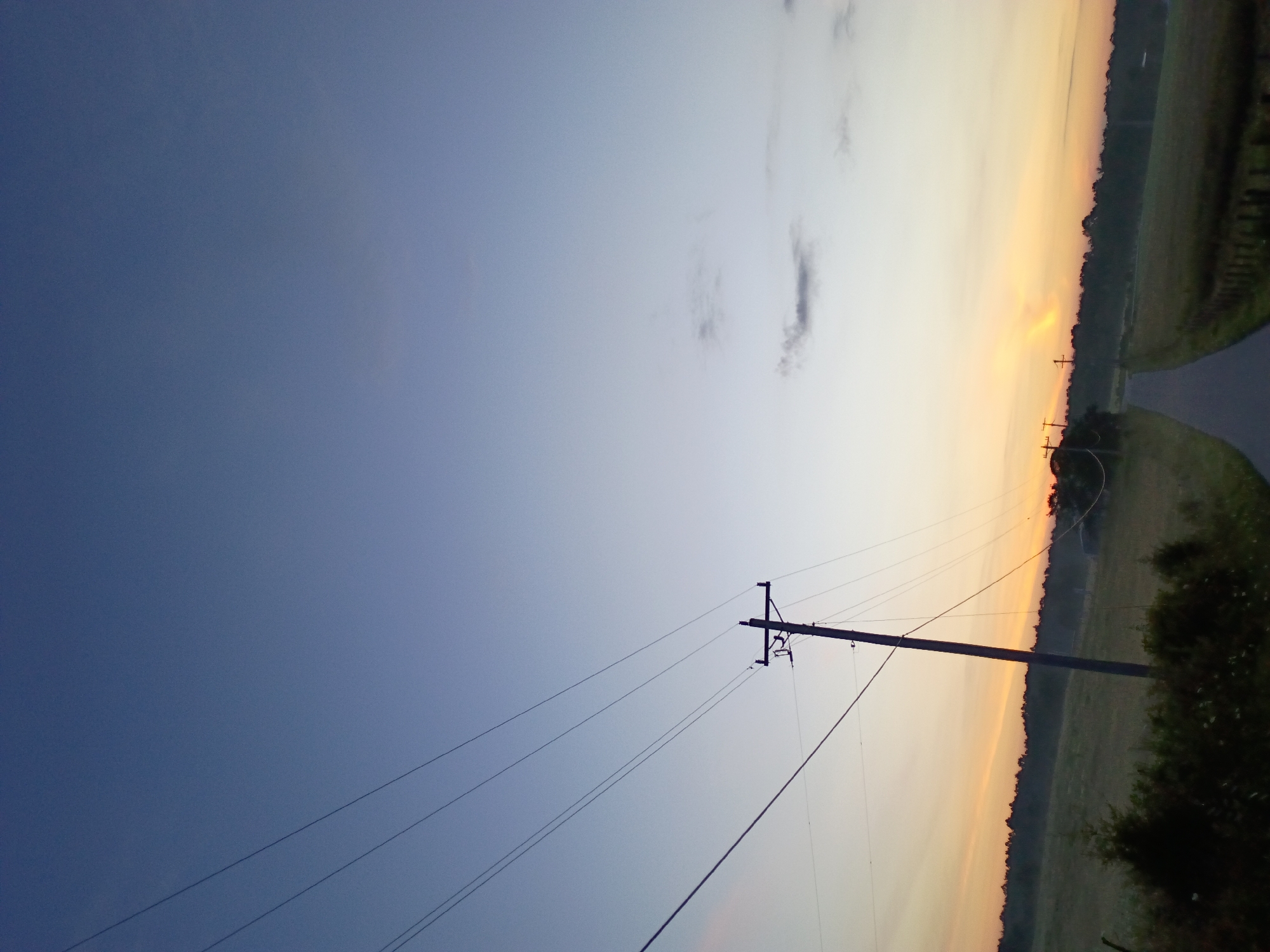 The image depicts an outdoor scene at dusk. A telephone pole with wires stretches diagonally across the frame, from the lower left to the upper right. The pole is silhouetted against a clear sky that transitions from a deep blue at the top to lighter shades of blue and orange near the horizon. A paved road curves into the distance, flanked by grassy fields and a line of trees. The lighting suggests it is either sunrise or sunset. There are no people or animals visible in the frame. The location is identified as being in Jeffersonville, United States.