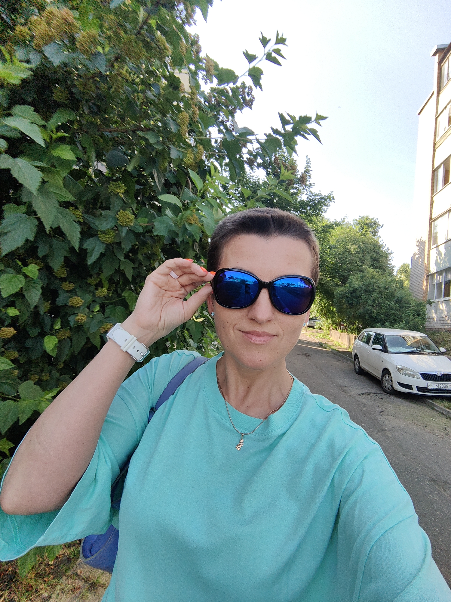 A woman stands outdoors in a city, holding her sunglasses with her right hand. She is wearing a turquoise t-shirt and a white smartwatch. Her hair is pulled back. Behind her, a white car is parked on the side of a street lined with trees and a brick building. The sky is clear and bright. The scene appears to be in Fanipal, Belarus.