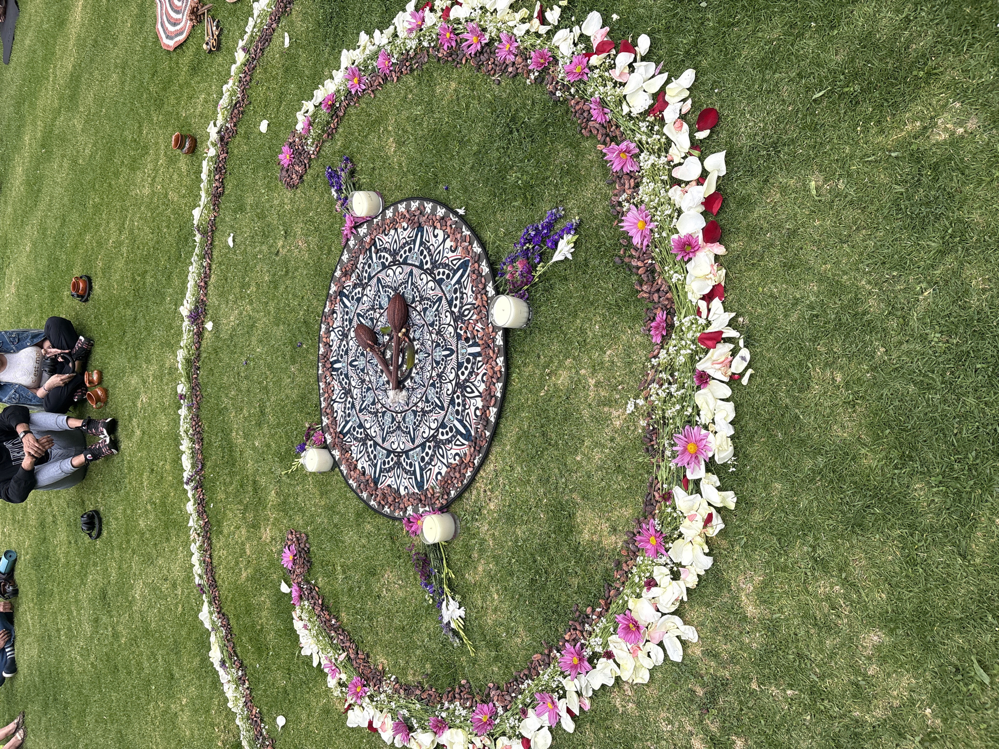 The image shows a circle of flowers and nuts on a green lawn in Santa Ana Acozautla, Mexico. The circle is incomplete and surrounds a patterned cloth on which three woody objects stand. Small bunches of flowers and candles are placed on the cloth's edge. People sit on the lawn near the circle.