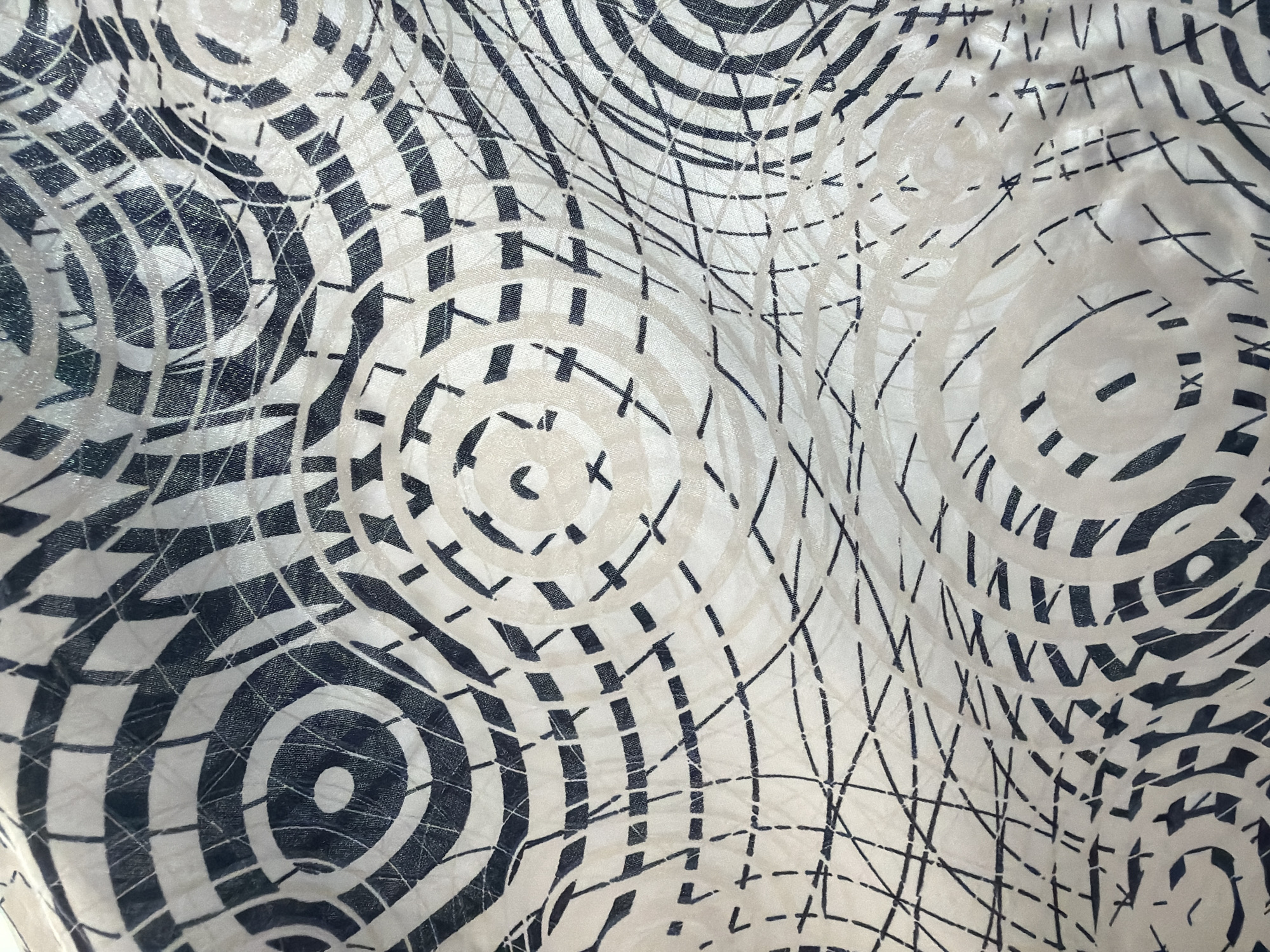 This media file is a close-up, top-down view of a fabric with a black and white abstract pattern. The pattern consists of concentric circles, some filled with solid black, others with a grid-like design of small squares, and some are outlined with thin black lines. There are also scattered thin black lines that crisscross the fabric, forming a net-like or chaotic scribble effect. The fabric itself appears to be textured, with visible woven threads and some slight creasing or rippling, indicating it is likely a garment or a piece of material. The lighting is even, with no strong shadows, suggesting an indoor setting or diffused natural light. There are no people, objects, or discernible scene components; the sole subject is the patterned fabric. No text is visible in the image. The context provided places this image in Dorsten, Germany, but there are no visual cues within the image to support or contradict this.