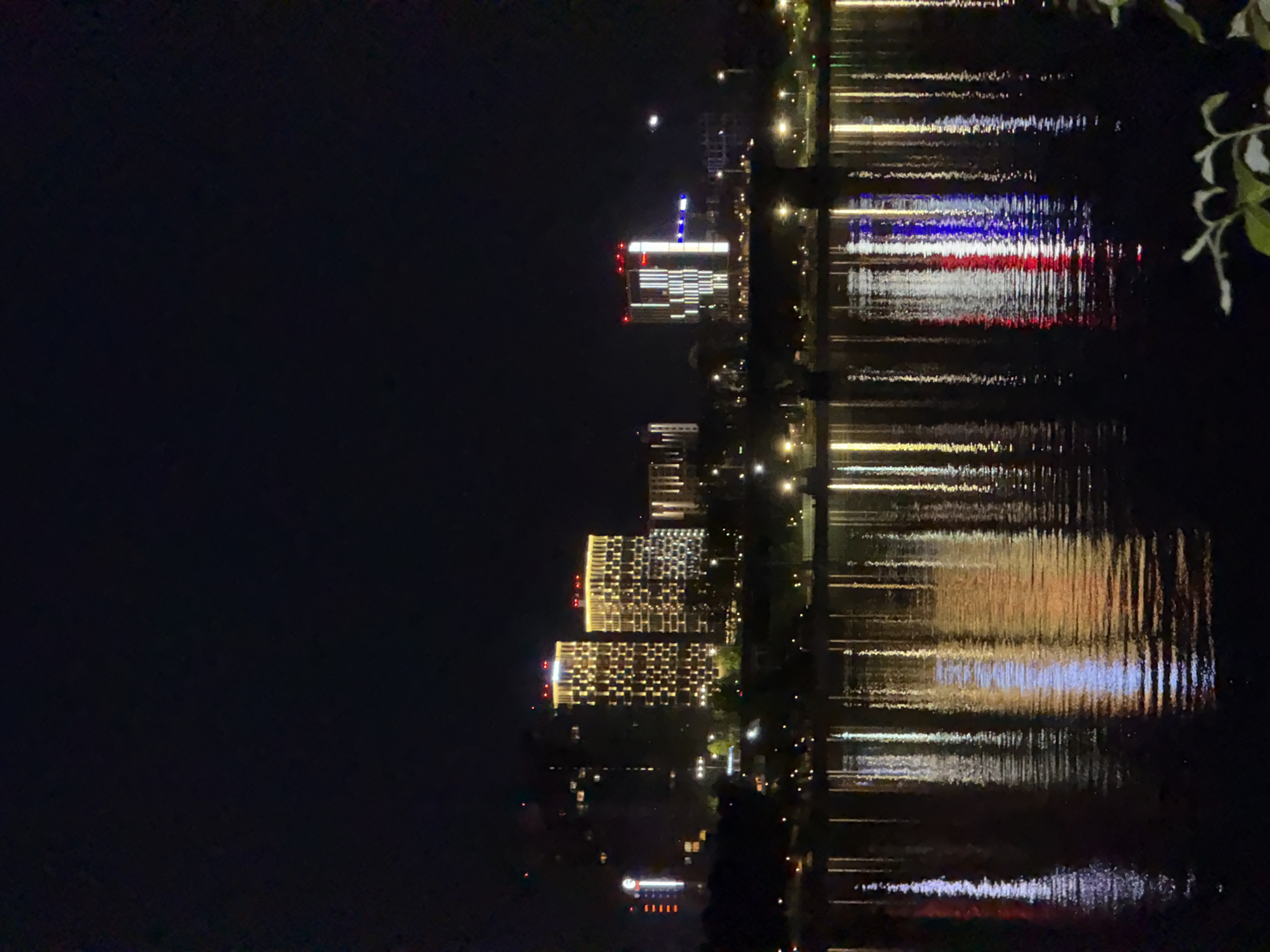 The image depicts a nighttime scene in Perm, Russia, showcasing the city skyline reflected on the Kama River. The architecture features several high-rise buildings illuminated with decorative lights. One building prominently displays red, white, and blue lights, resembling the colors of the Russian flag. Other buildings are lit up with a pattern of warm, yellowish lights. The dark water of the river mirrors these lights, creating a shimmering effect. A branch with leaves is partially visible on the right side of the frame, suggesting the photo was taken from a riverside location. The rest of the sky is dark, indicating it is nighttime.