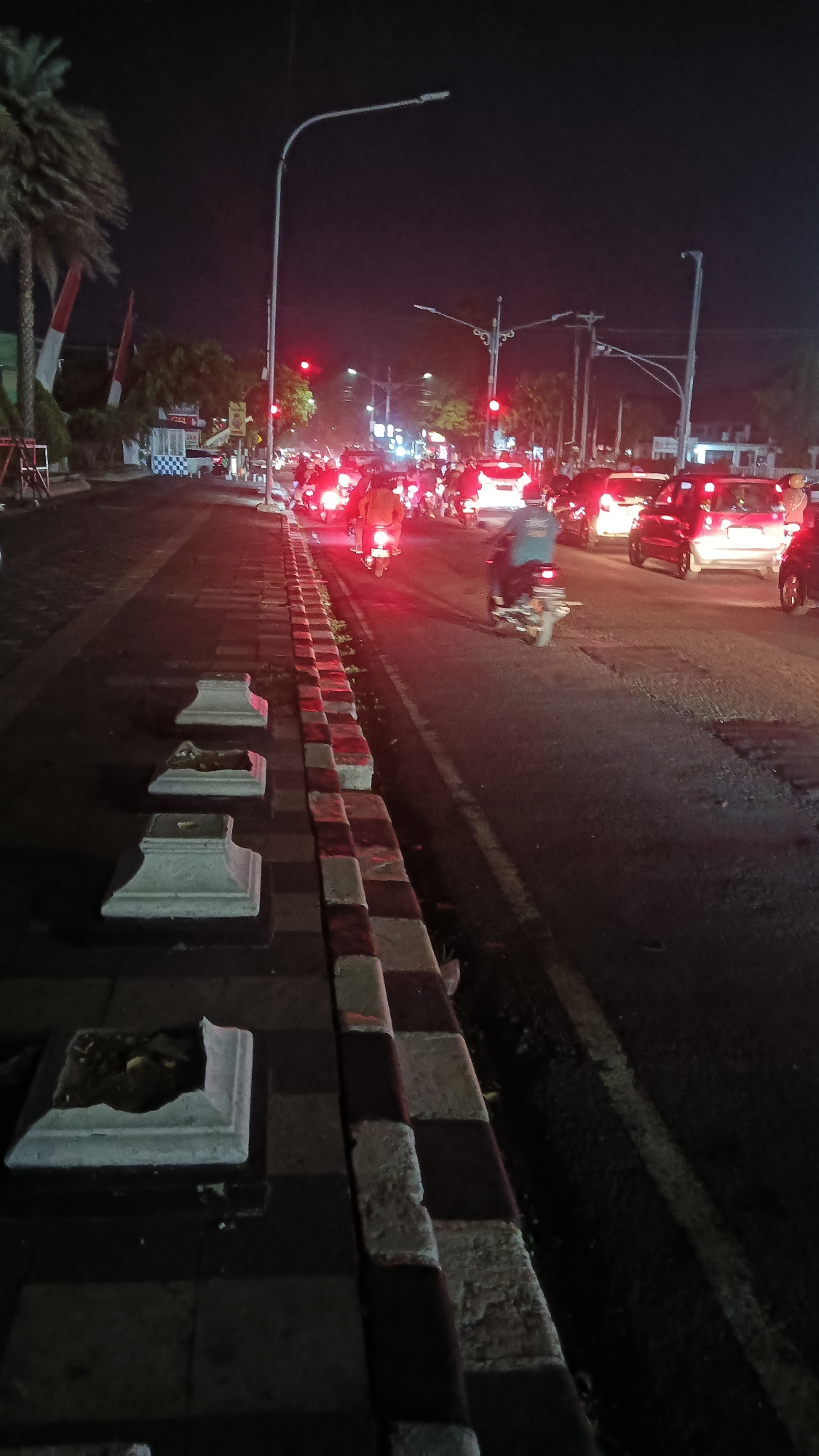 This image depicts a nighttime street scene in Sragen, Indonesia, likely on a busy road due to the presence of a traffic jam. The foreground features a sidewalk with decorative concrete planters, some of which appear damaged. The sidewalk is bordered by a checkerboard pattern of light and dark stones. The street itself is filled with numerous motorcycles and cars, many with their headlights and taillights illuminated, indicating a traffic backup. Red traffic lights are visible, contributing to the congestion. Streetlights illuminate the scene, casting light and shadows. The background shows more traffic, buildings, and trees, with a palm tree visible on the left side of the image. The overall impression is that of a typical urban street at night experiencing moderate traffic.