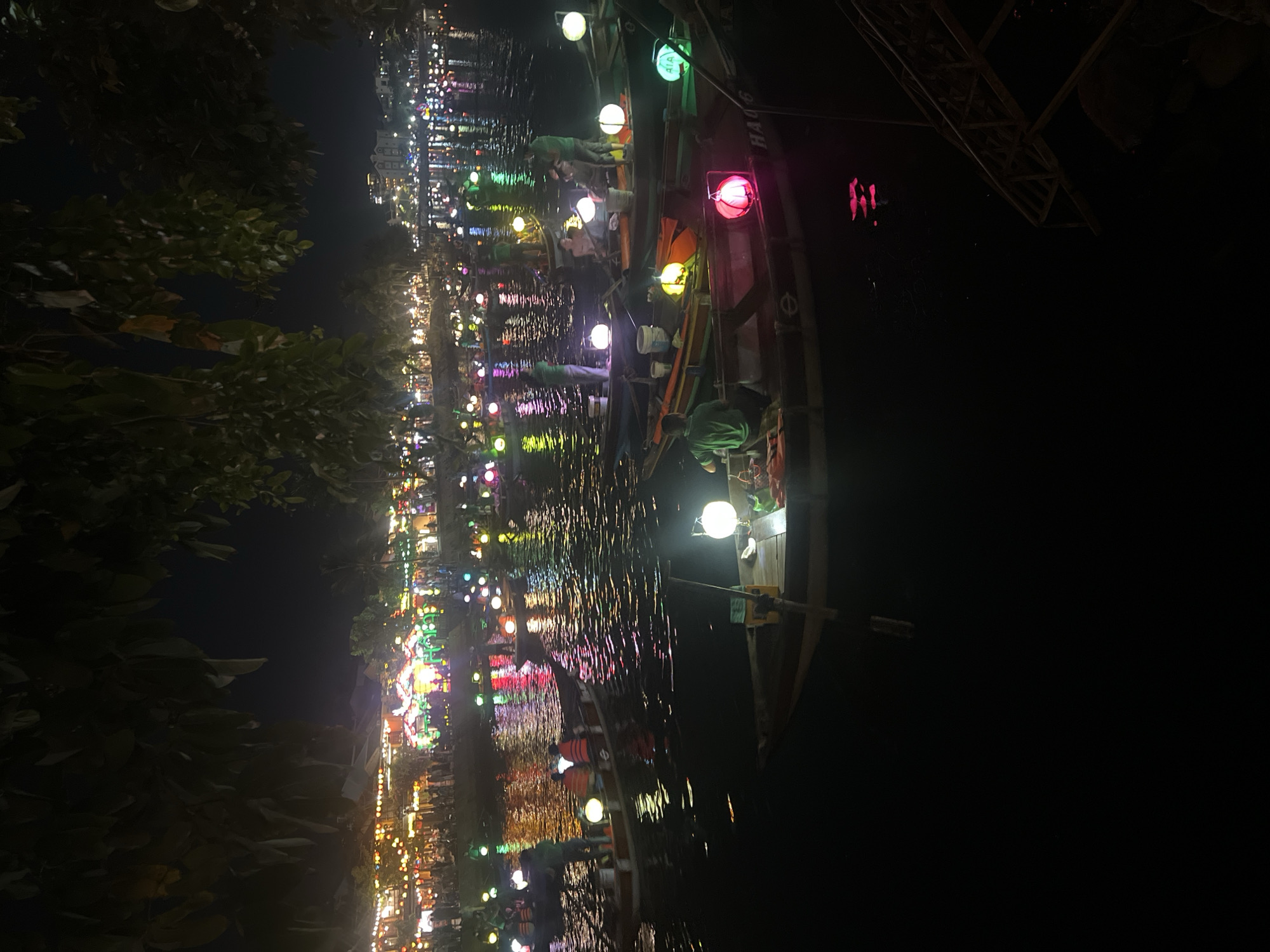 The image depicts a nighttime scene along the Thu Bon River in Hoi An, Vietnam. The river is filled with numerous small wooden boats, many of which are adorned with colorful illuminated lanterns, casting reflections on the dark water. Several people, likely boat operators and tourists, are visible in and around the boats. The background shows a vibrant waterfront with buildings illuminated by strings of lights, suggesting a lively marketplace or tourist area. The overall atmosphere is festive and tranquil, typical of Hoi An's nighttime ambiance.