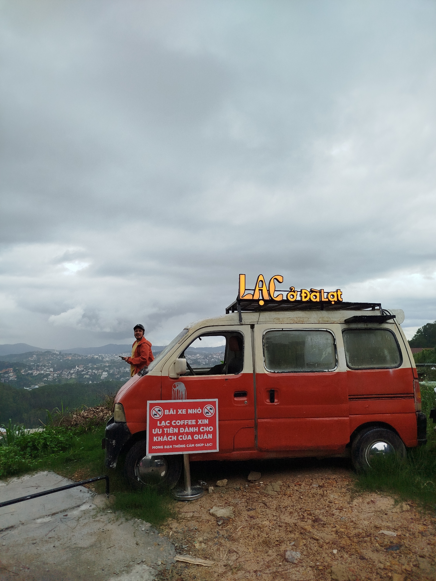The image depicts a scene in Xuan Truong - Da Lat, Vietnam. A two-toned, orange and white van, branded "LẠC ở ĐÀ LẠT" with illuminated letters on its roof rack, stands on a dirt and grassy patch. A sign on the van reads "BÃI XE NHỎ LẠC COFFEE XIN ƯU TIÊN DÀNH CHO KHÁCH CỦA QUÁN MONG BẠN THÔNG CẢM GIÚP LẠC!" (Small parking lot. LẠC COFFEE prioritizes customers of the shop. We ask for your understanding in helping LẠC!). A man in an orange jacket stands adjacent to the van, looking at the camera. In the background, a cityscape stretches across the valley under a cloudy sky.