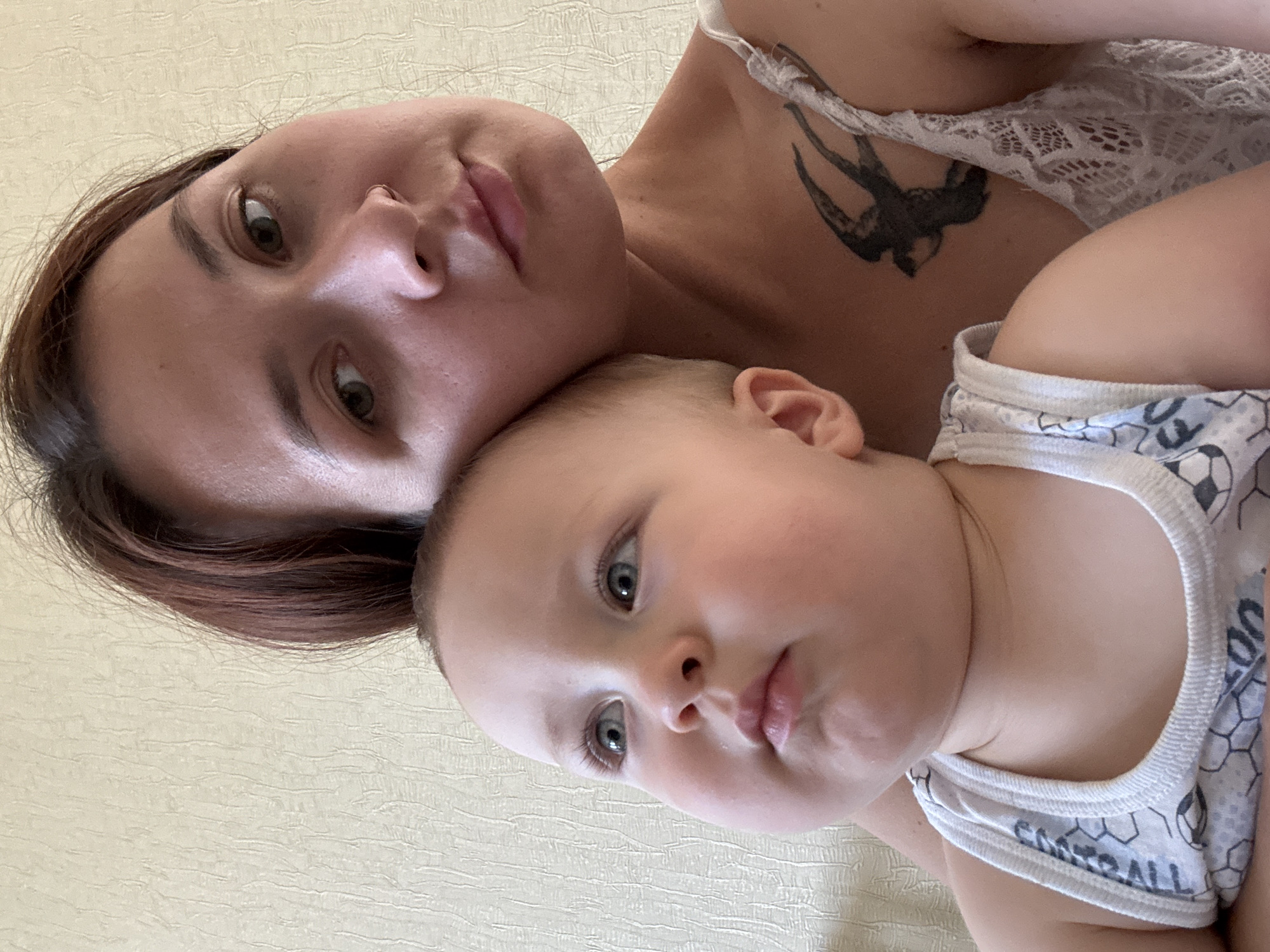 The image shows a close-up selfie of a woman and a baby. The woman has brown hair and is wearing a white lace top. She has a tattoo of a bird on her chest. The baby is fair-skinned with blue eyes and is wearing a white tank top with a soccer ball pattern. The background appears to be a textured, light-colored wall. The lighting suggests an indoor setting, possibly a home. The specific location in Naberezhnye Chelny, Russia, cannot be determined from the image itself.